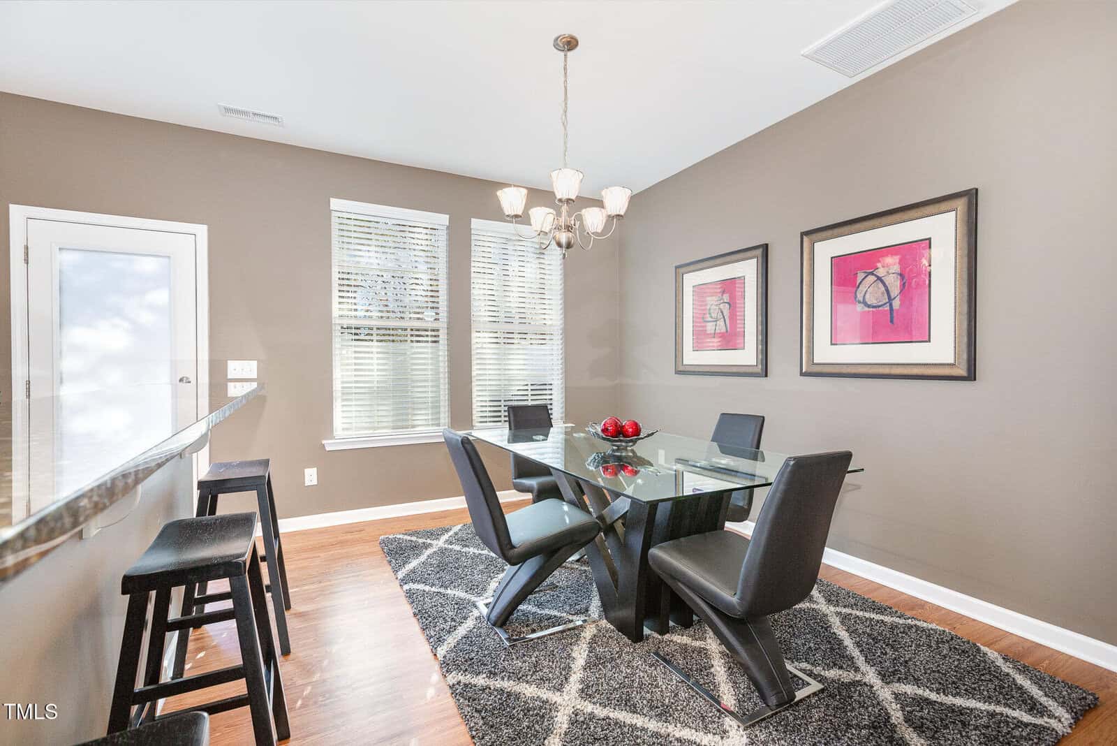 Modern dining area with a glass table, four black chairs, a patterned rug, two framed artworks on taupe walls, and a trio of barstools by a counter. Large windows let in natural light.