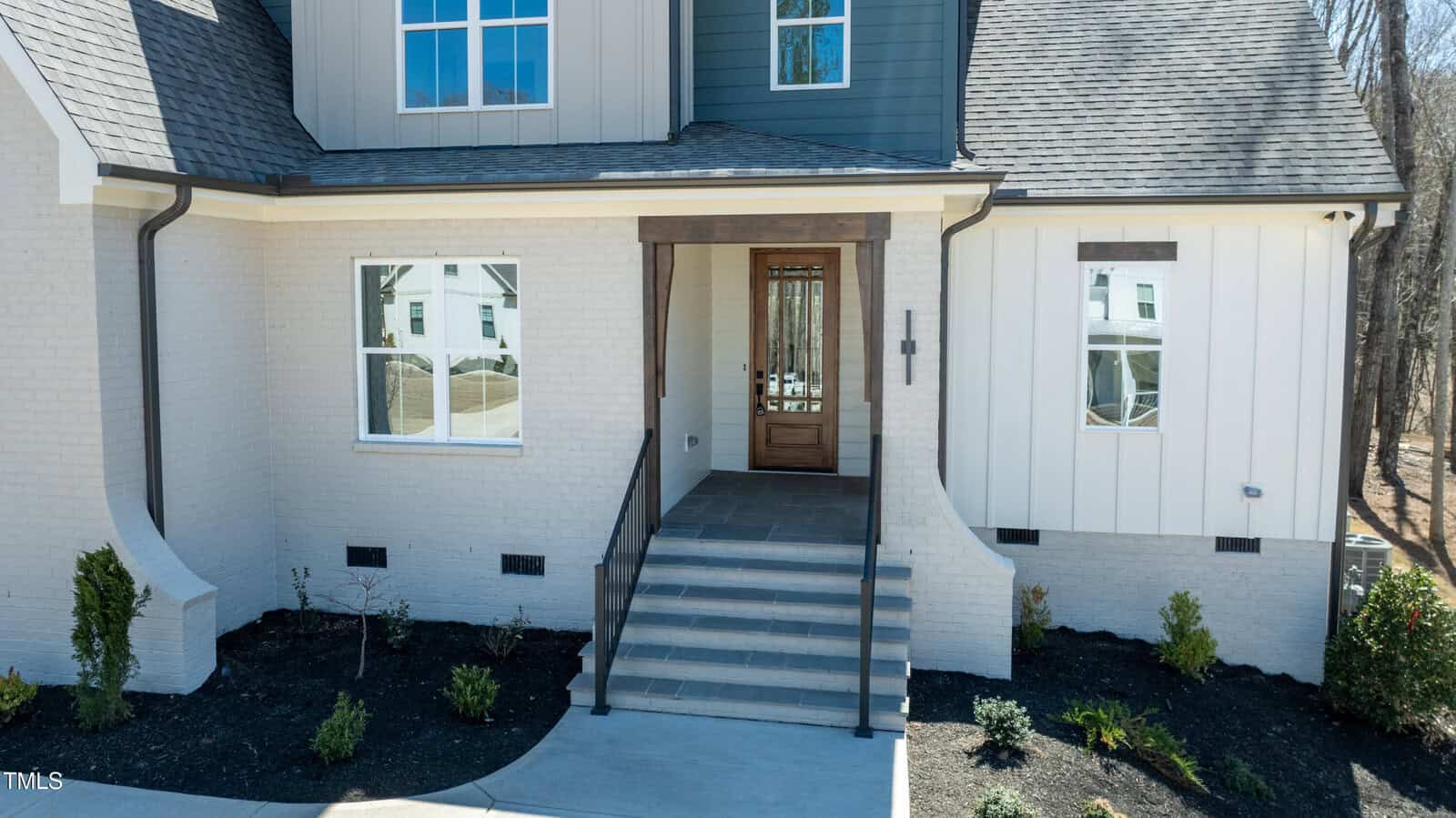 Front entrance of a modern house with light brick and vertical siding, wooden trim, a glass-paneled front door, steps leading up to the porch, and minimal landscaping with small bushes.