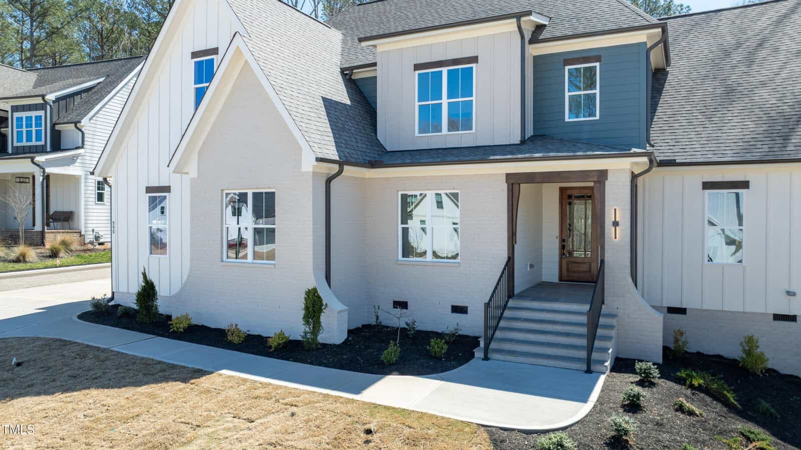 Modern two-story house with light brick and siding exterior, large windows, a wooden front door with glass panels, and a landscaped yard with newly planted shrubs and a concrete walkway.