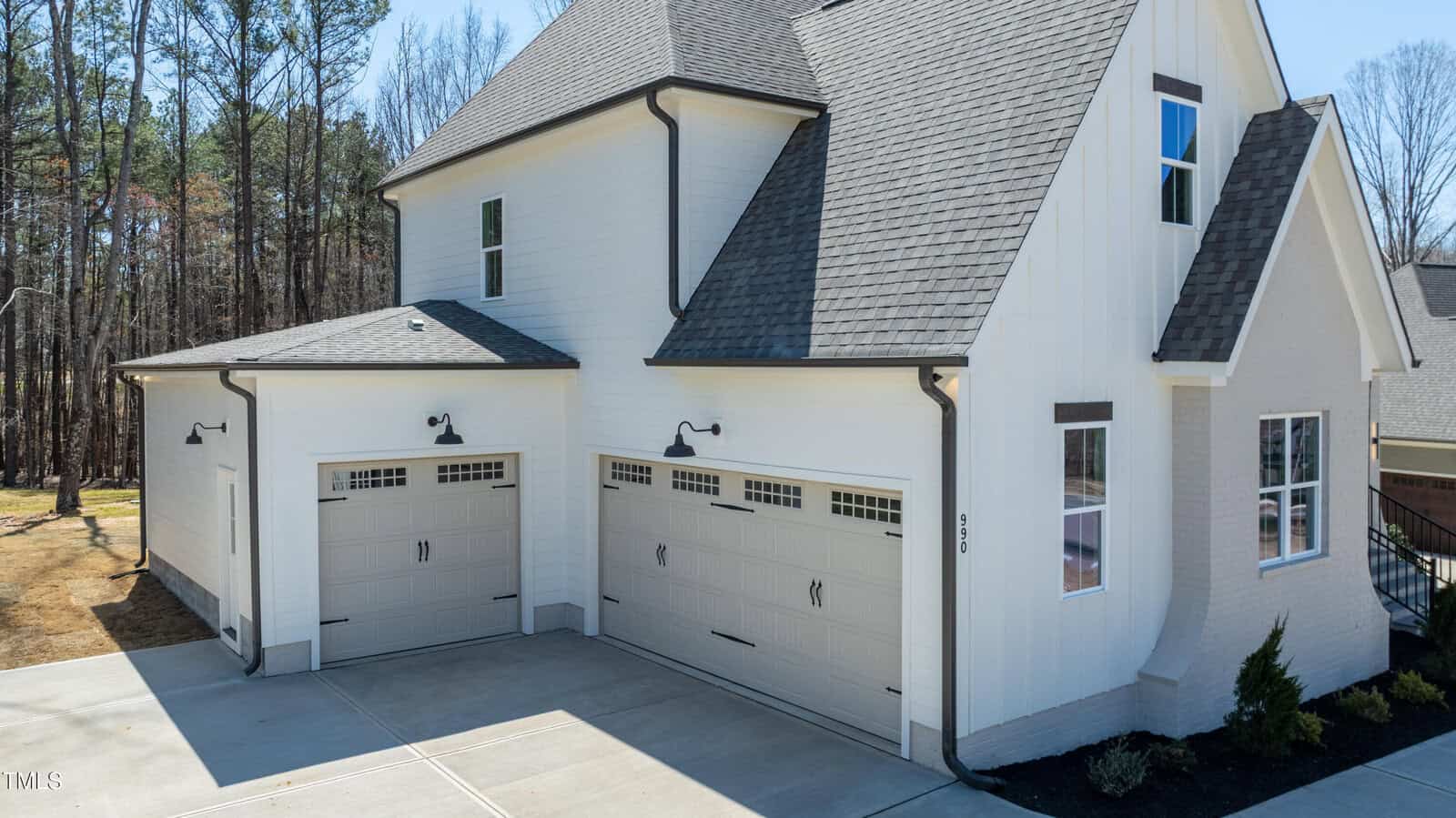 A modern white house with gray roof shingles, featuring three large garage doors, black exterior lights, and a concrete driveway. Tall trees are visible in the background.