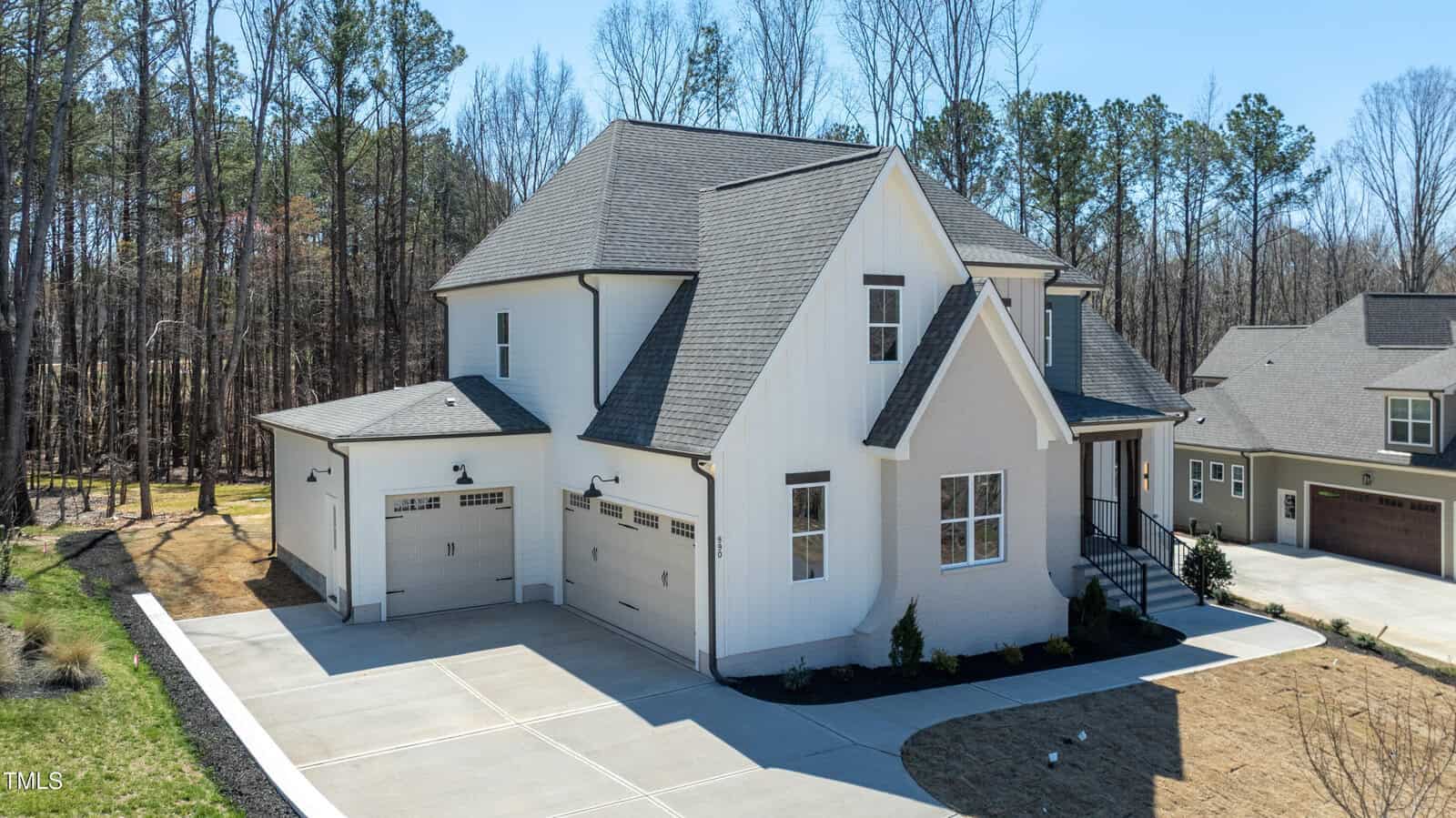 A modern two-story white house with gray roofing, three garage doors, and large windows, surrounded by trees and set on a spacious concrete driveway in a suburban neighborhood.