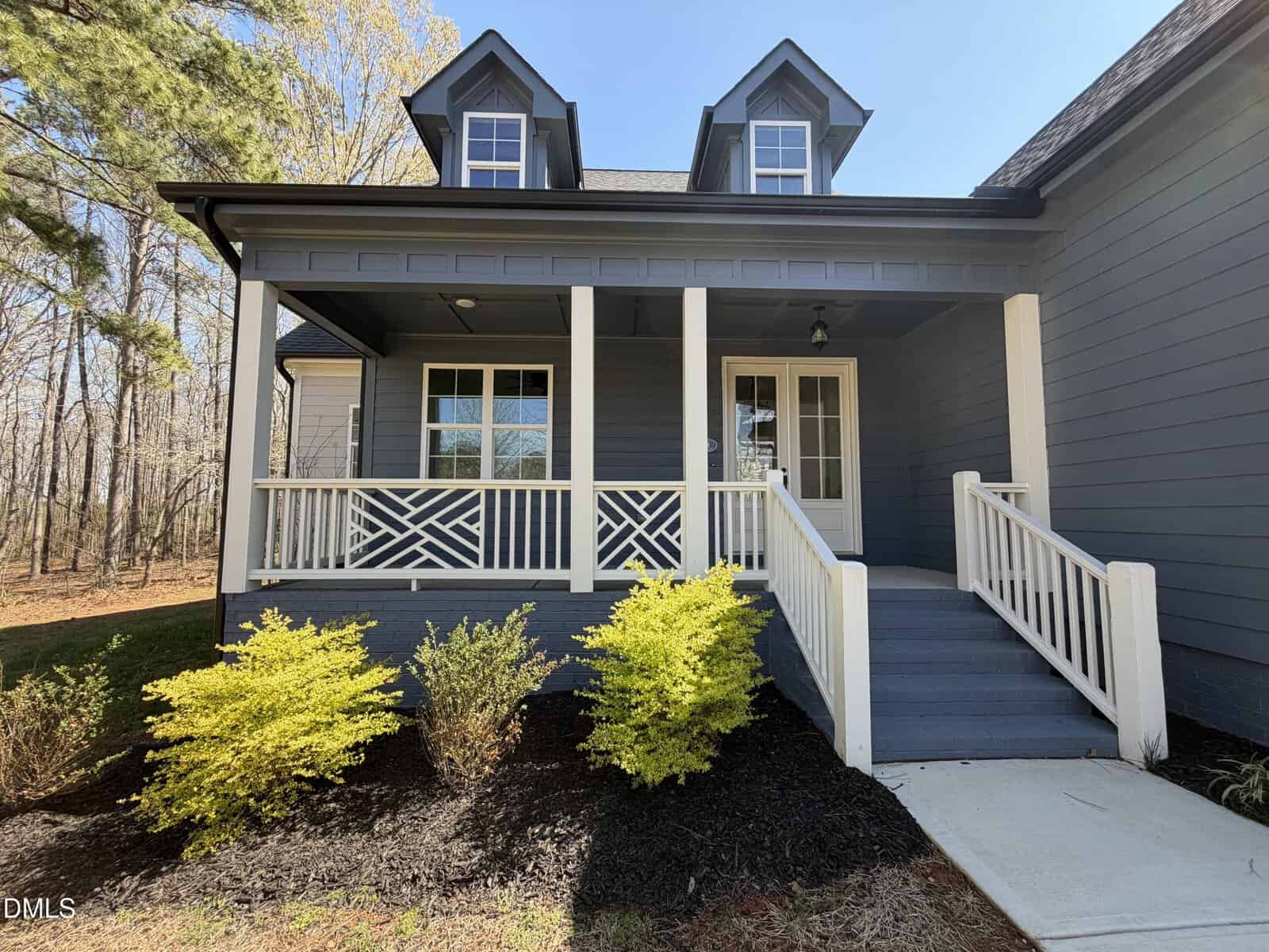 A front view of a house with blue siding, a covered porch with white railings, two dormer windows, and a ramp leading to the entrance. Yellow-green shrubs are planted in dark mulch beside the porch.