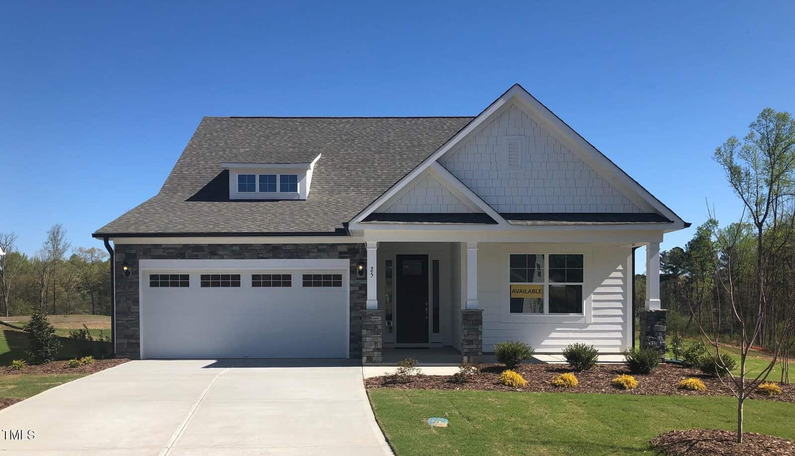 Single-story suburban house with white siding, gray stone accents, a covered front porch, attached two-car garage, manicured lawn, and a sign in the front window reading "Available" on a clear, sunny day.