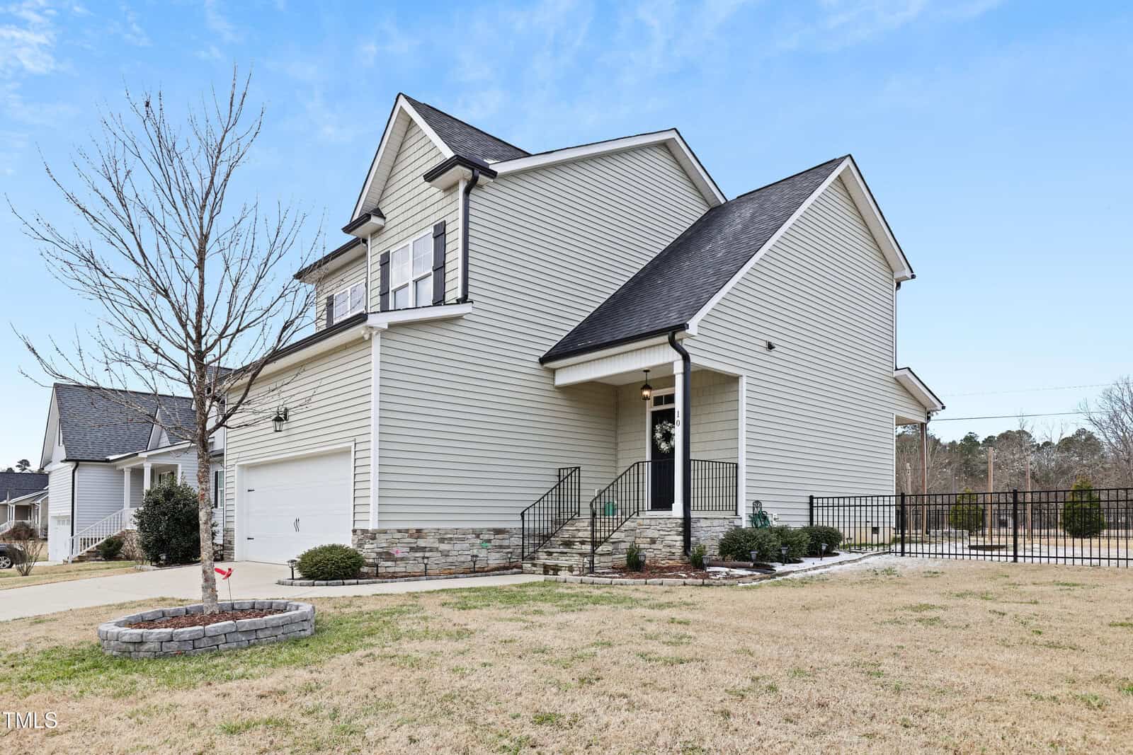 A modern two-story house with gray siding, white trim, and a black roof. The home has a front porch, a two-car garage, a small landscaped area, and a fenced backyard. The sky is clear and the lawn is brown.