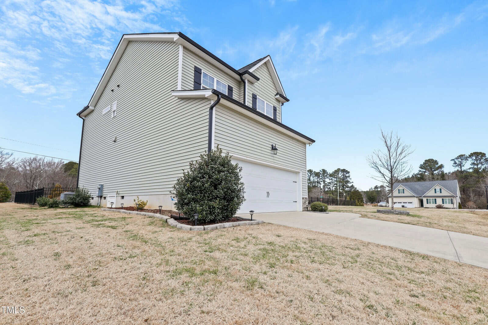 Two-story suburban house with beige siding, white trim, and a double garage. The yard features trimmed bushes and dry grass, with a concrete driveway leading to the garage. Another house is visible in the background.