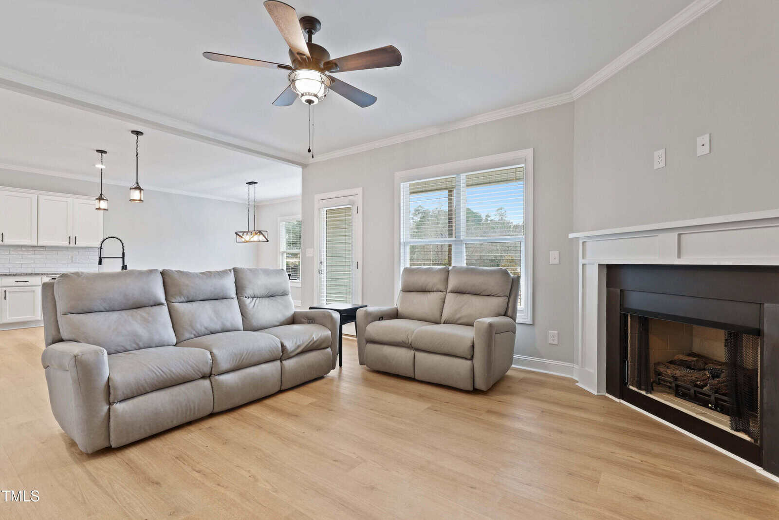 A modern living room with light wood floors, gray reclining sofa and chair, ceiling fan, fireplace, and large windows. The space opens to a white kitchen with pendant lights and a small dining area.