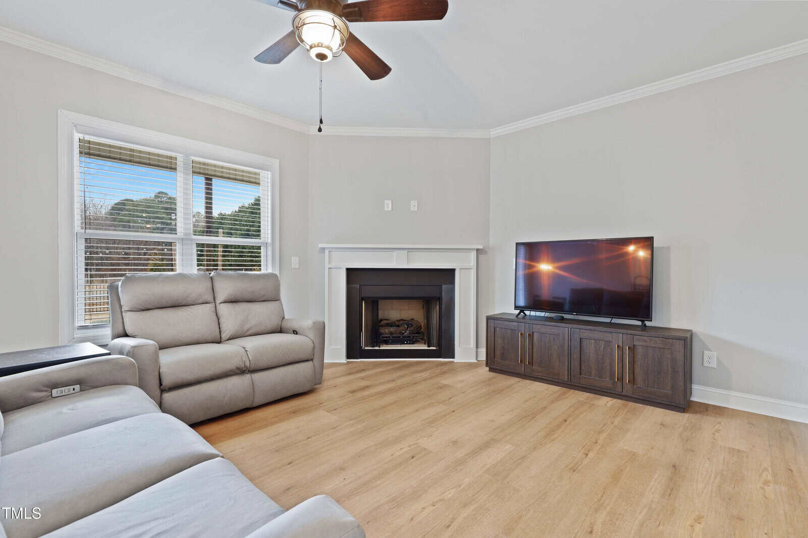 A living room with light wood flooring, two gray recliner sofas, a fireplace with a white mantle, a flat-screen TV on a dark cabinet, and a ceiling fan with light. Sunlight enters through large windows on the left.