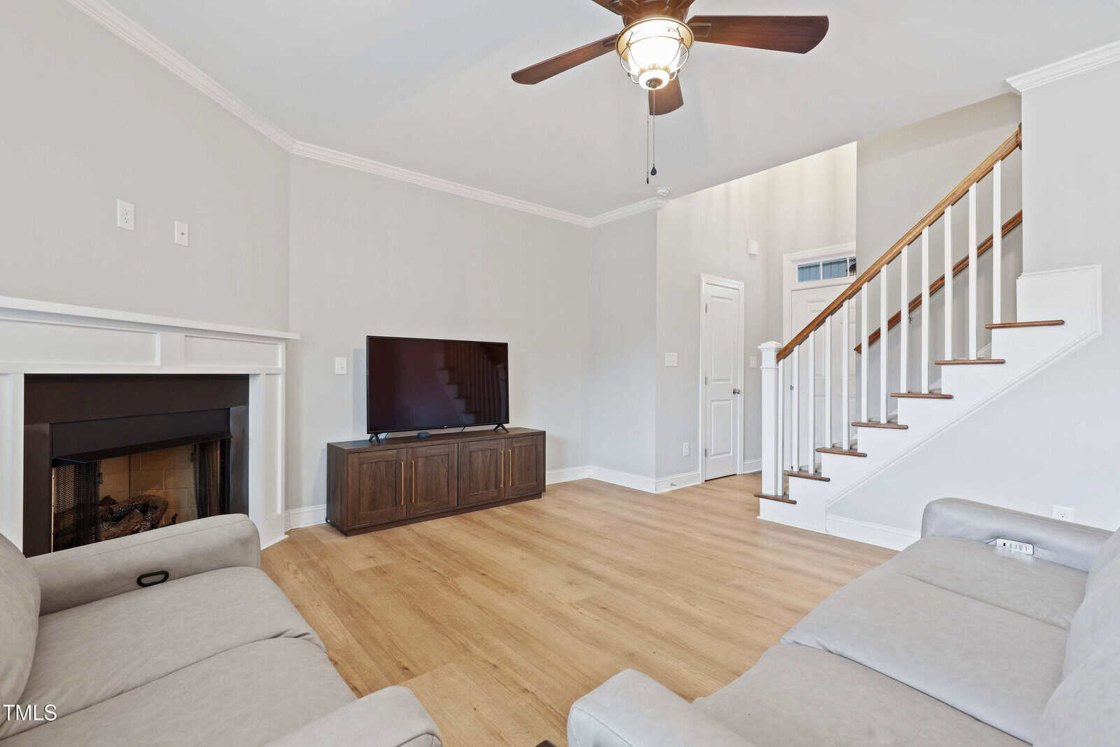 A living room with light wood flooring, light gray walls, a ceiling fan with light, a wall-mounted TV on a dark media console, a fireplace, two beige sofas, and a staircase with white spindles and wood handrail.
