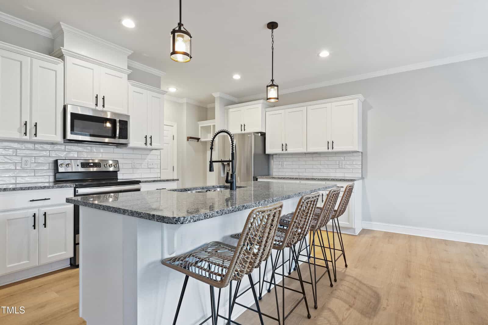 Modern kitchen with white cabinets, stainless steel appliances, a gray granite island with black faucet, and four woven barstools. Light wood floor, subway tile backsplash, and pendant lights above the island.