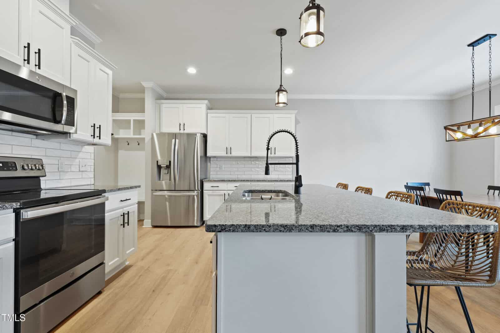 Modern kitchen with white cabinets, stainless steel appliances, a granite island with a black faucet, wooden floor, and wicker barstools. A dining area with a chandelier is visible in the background.