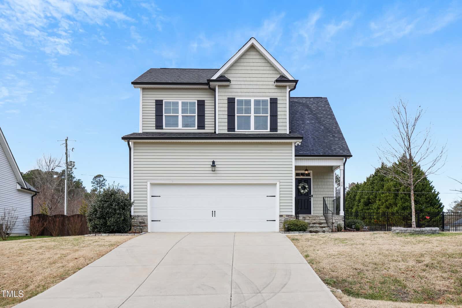 A two-story suburban house with gray siding, black shutters, a covered front door, and a double garage. The driveway and lawn are in front, with trees and a partly cloudy sky in the background.