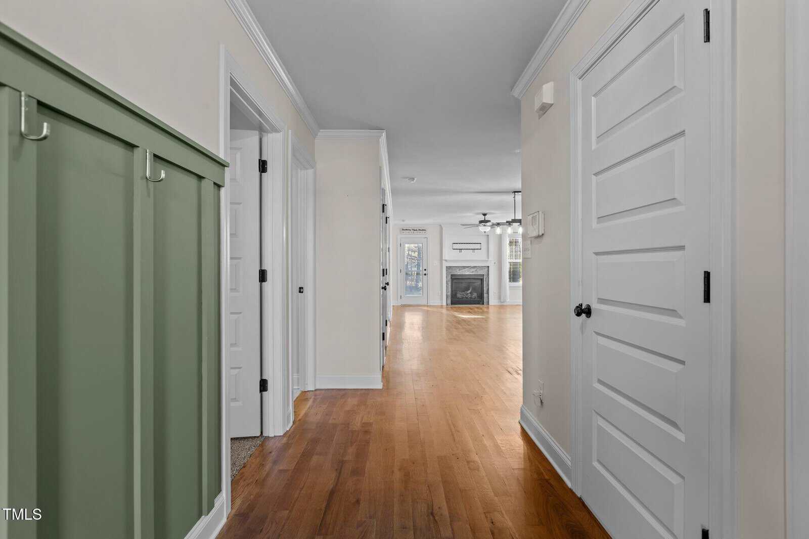 A hallway with light wood floors, white doors and trim, green wall paneling with coat hooks on the left, leading to a living room with a fireplace and ceiling fan in the background.