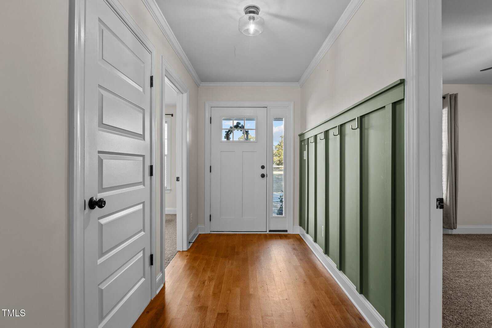A hallway with light walls, wooden flooring, white doors, a white front door with glass panels, and a green board-and-batten accent wall with coat hooks. Natural light comes in through the glass.