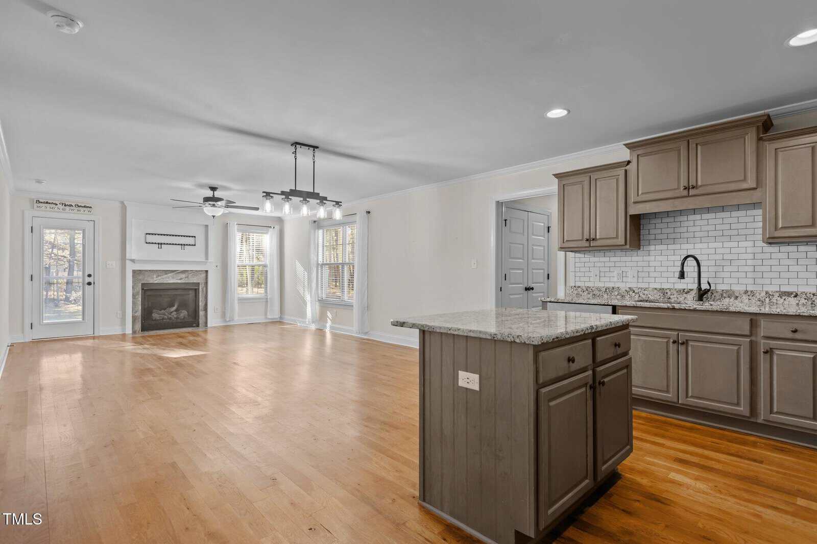 Open-concept living area with light wood flooring, a kitchen with gray cabinets and a center island, white subway tile backsplash, granite countertops, and a living space with a fireplace and large windows.