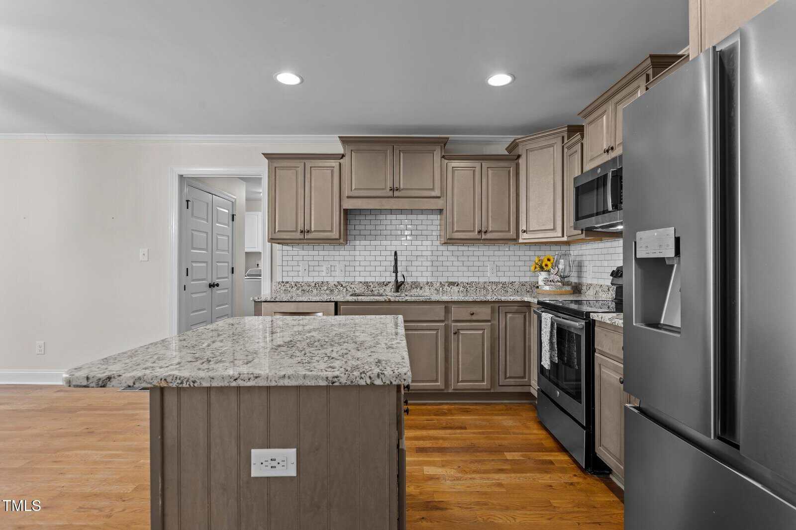 Modern kitchen with light brown cabinets, granite countertops, white subway tile backsplash, stainless steel appliances, an island with built-in outlet, and wooden flooring. A vase with yellow flowers sits by the stove.
