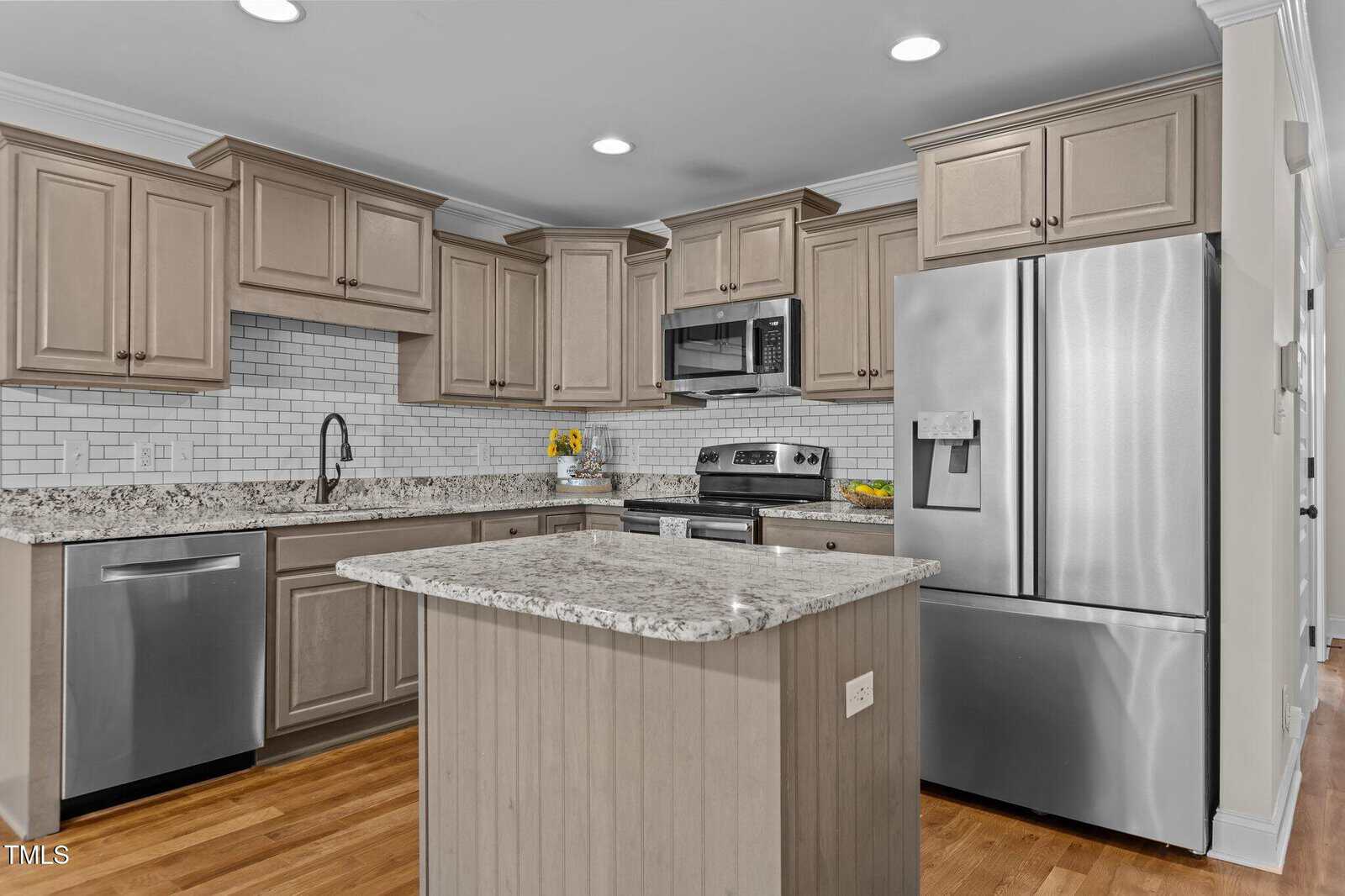 Modern kitchen with light wood cabinets, stainless steel appliances, a granite-topped island, subway tile backsplash, dishwasher, and wooden flooring. A bowl of lemons and a yellow flower arrangement are visible on the counters.