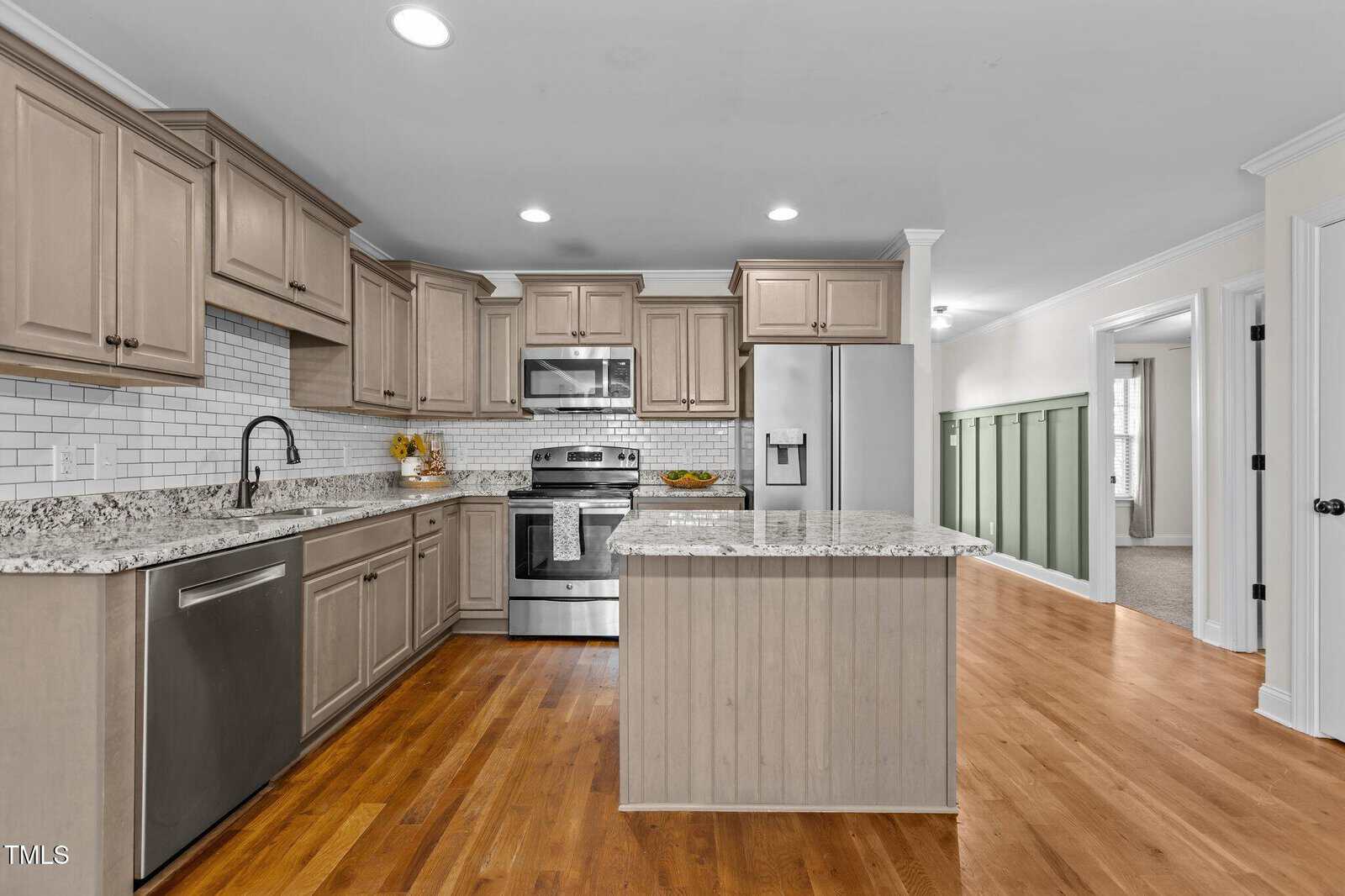 Kitchen with light wood cabinets, stainless steel appliances, granite countertops, white subway tile backsplash, an island, and hardwood floors. A hallway with green wainscoting is visible to the right.