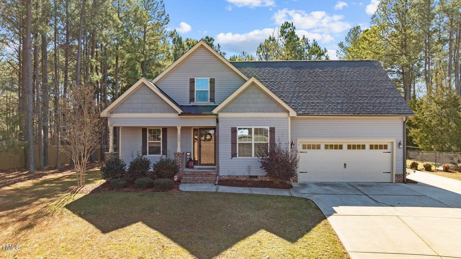 A single-story house with beige siding, gray roofing, and a two-car garage. The front yard has bushes and a tree, and a concrete driveway leads to the garage. Tall pine trees are visible in the background.