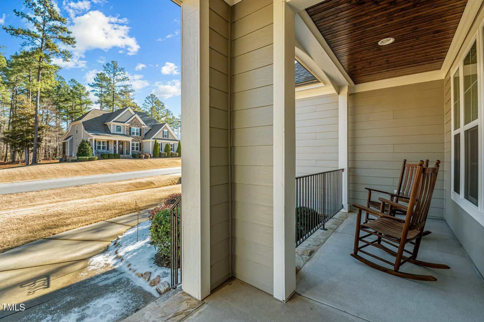 A covered front porch with two wooden rocking chairs overlooks a suburban street lined with large houses, trees, and a clear blue sky.