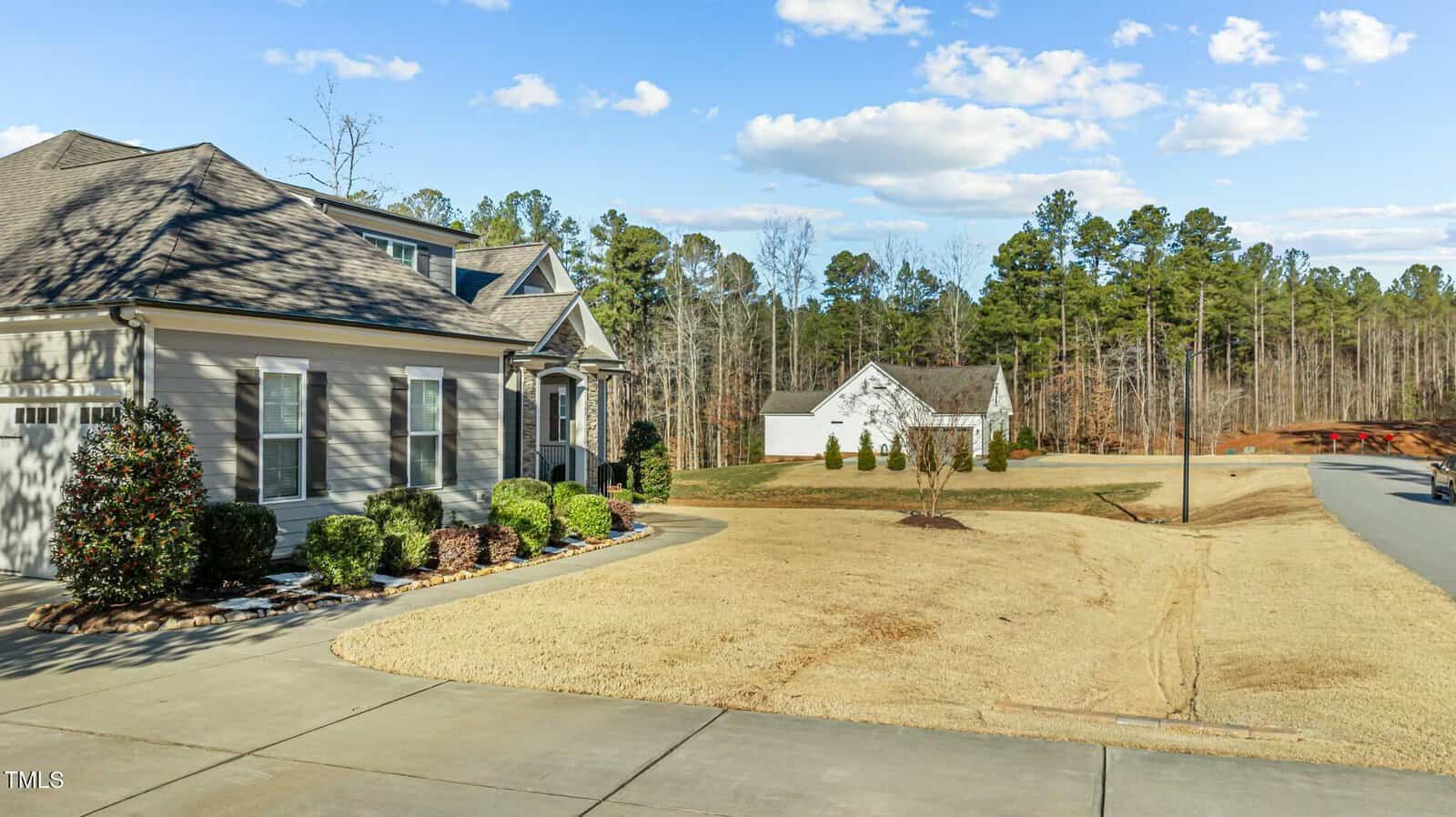 A suburban home with light-colored siding and a manicured front yard sits beside a paved driveway. In the background, there is a detached garage and a wooded area with tall trees under a blue sky.