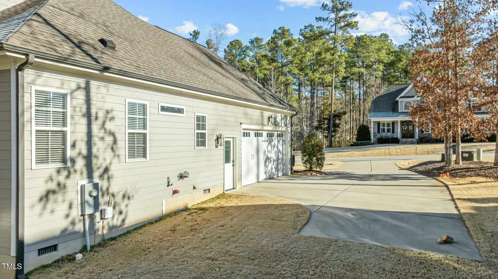 A light-colored house with a two-car garage and a long driveway, surrounded by brown grass and trees. Another house is visible in the background across the street.