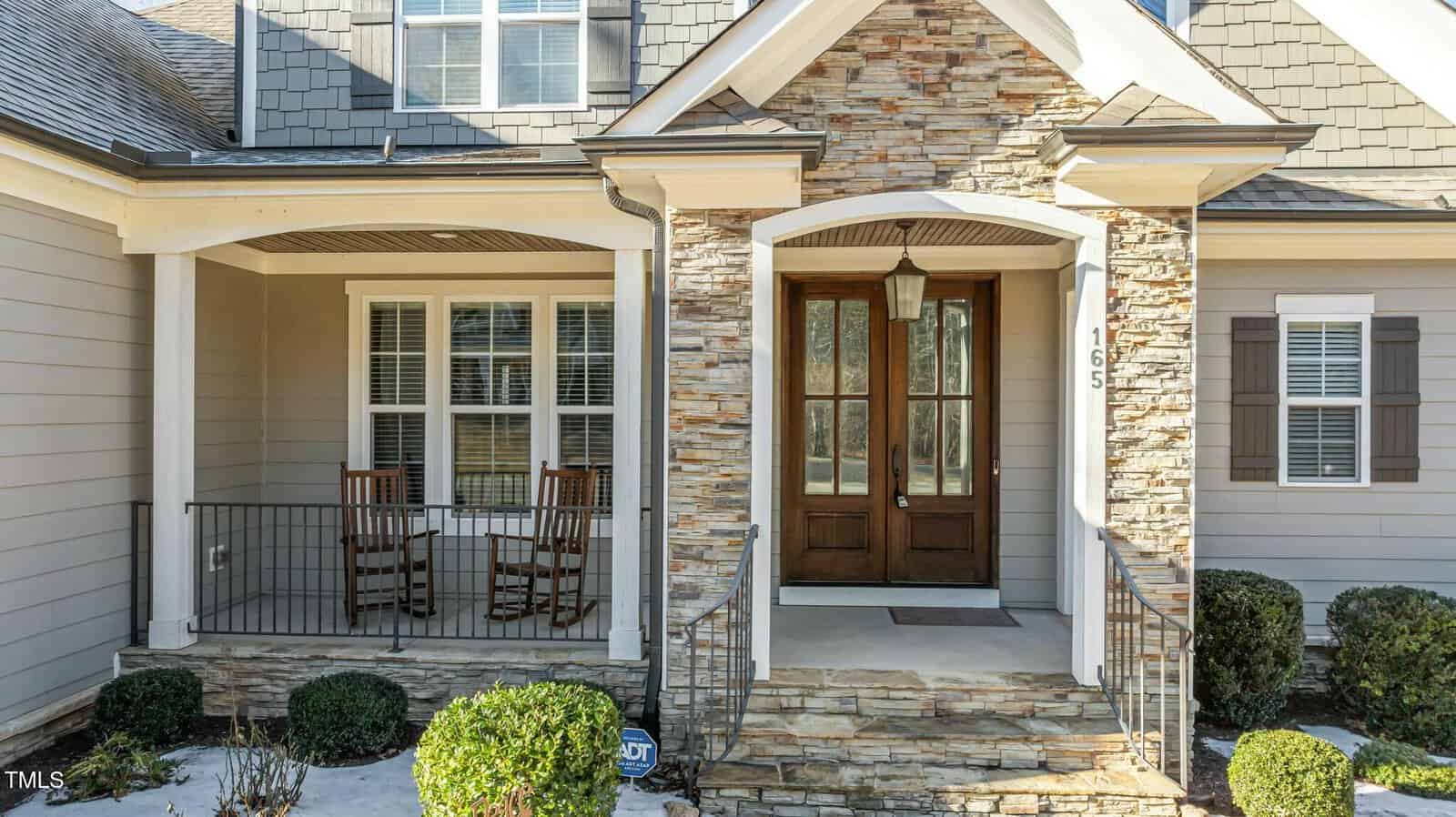 Front view of a house with stone and siding exterior, a covered porch with two wooden rocking chairs, white-trimmed windows, a double wooden front door, and neatly trimmed bushes along the walkway.