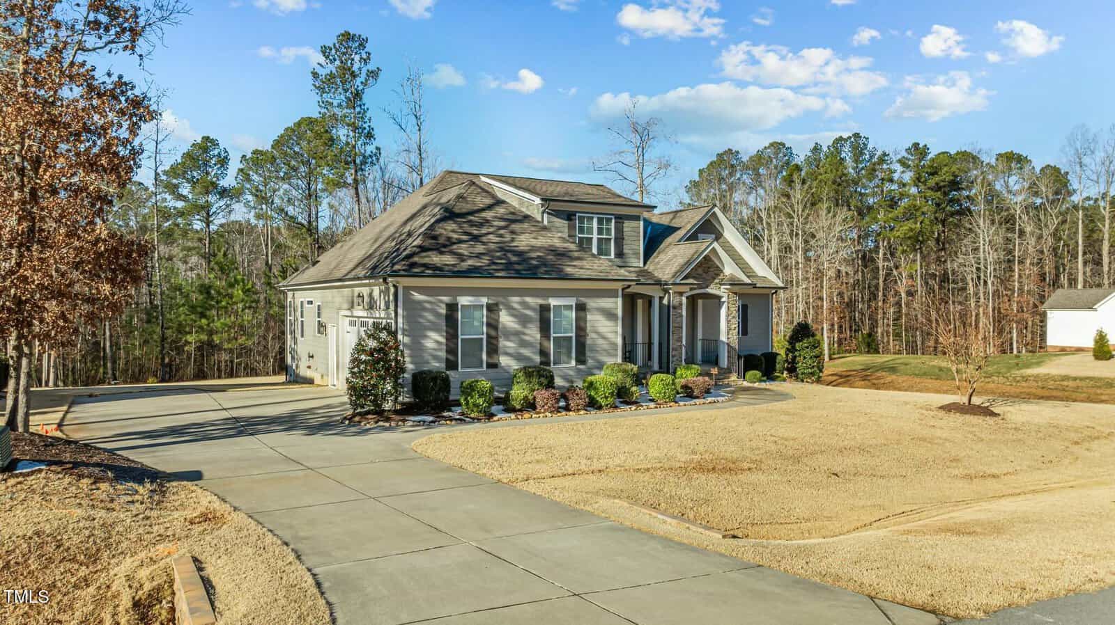 Single-story house with gray siding and dark roof, surrounded by sparse landscaping and a curved driveway, set against a backdrop of trees under a blue sky with scattered clouds.