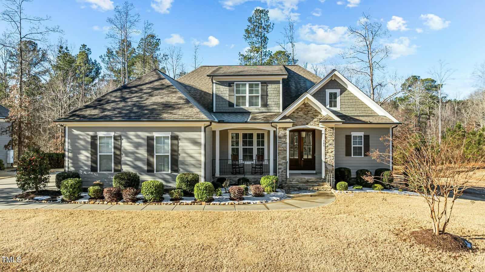 A single-story house with gray siding, white trim, and a covered front porch. The yard features trimmed bushes, a stone walkway, and dry grass. Trees and a clear sky are in the background.