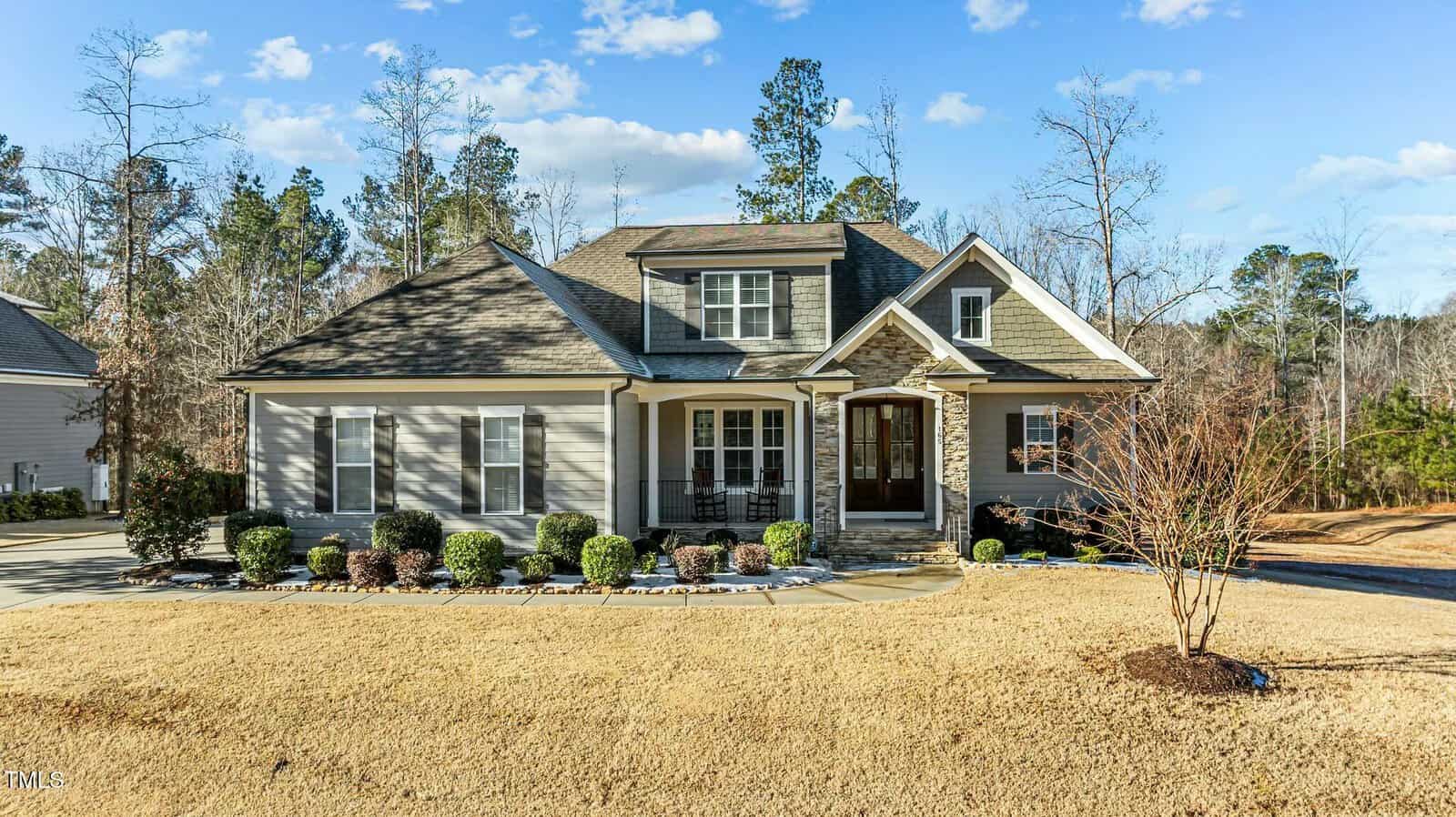 Single-story house with gray siding, stone accents, and a front porch with double doors. The yard has trimmed shrubs, a small tree, and dry, yellow grass. Trees and blue sky are in the background.