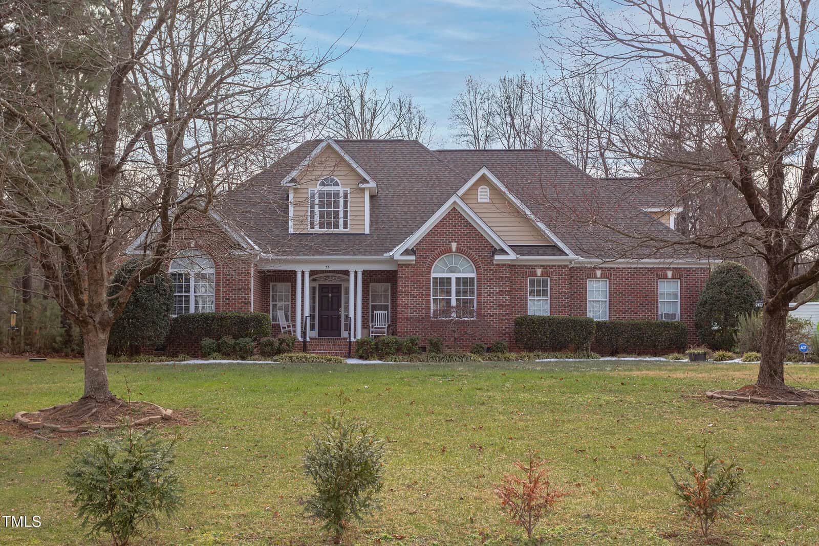 A two-story brick house with white trim, multiple gables, and a covered front porch. The front yard has green grass, small shrubs, and several large leafless trees.