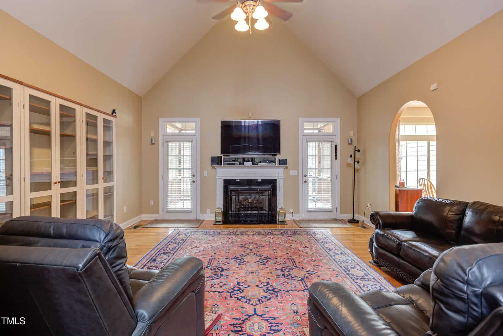 A living room with vaulted ceiling, two black leather sofas and two recliners, a fireplace with a mounted TV above it, built-in shelves on the left, a patterned rug, and hardwood floors.