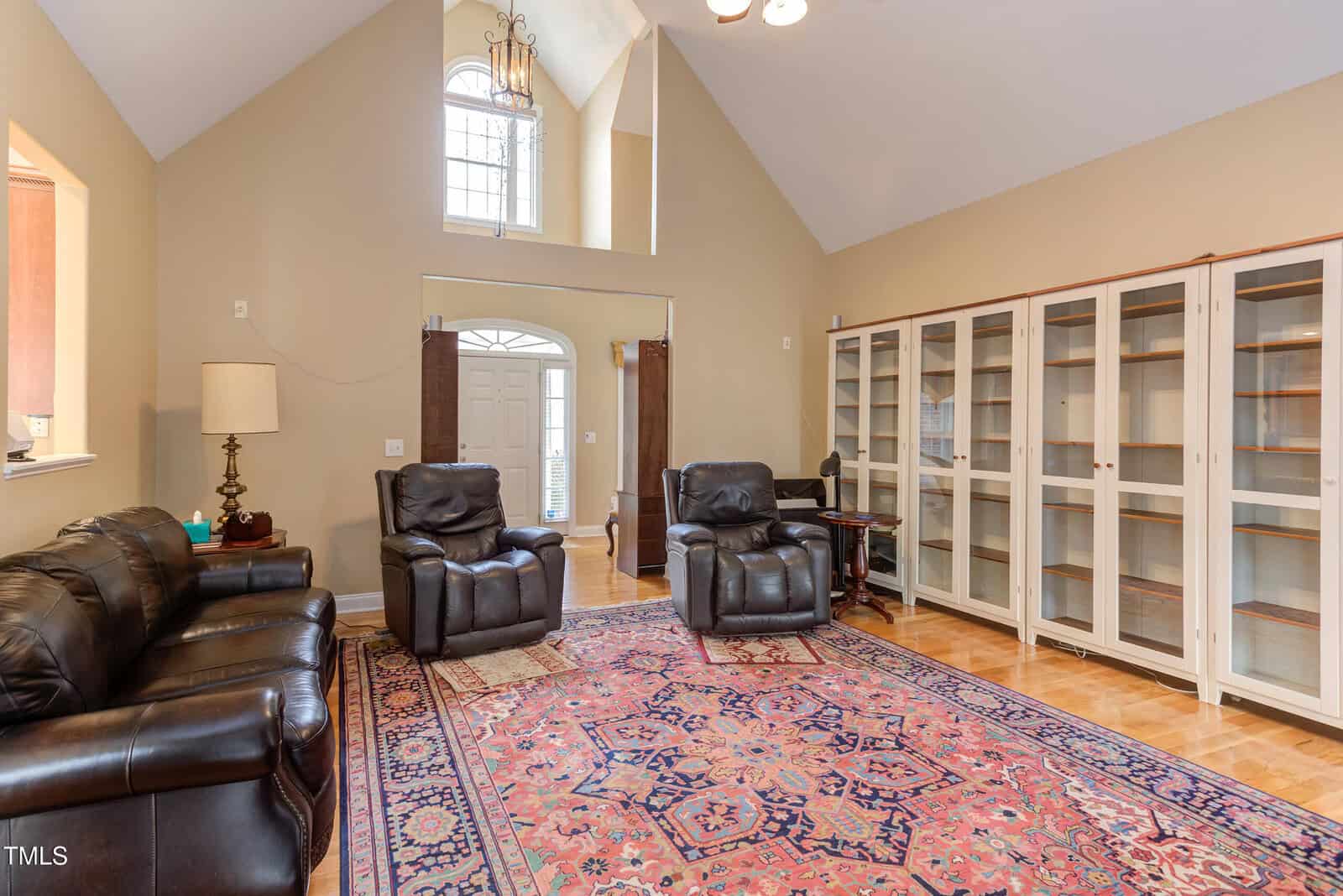 A living room with leather couches and recliners, a large patterned rug, glass-front bookshelves along one wall, and a high vaulted ceiling with a hanging light fixture near tall windows by the entrance.
