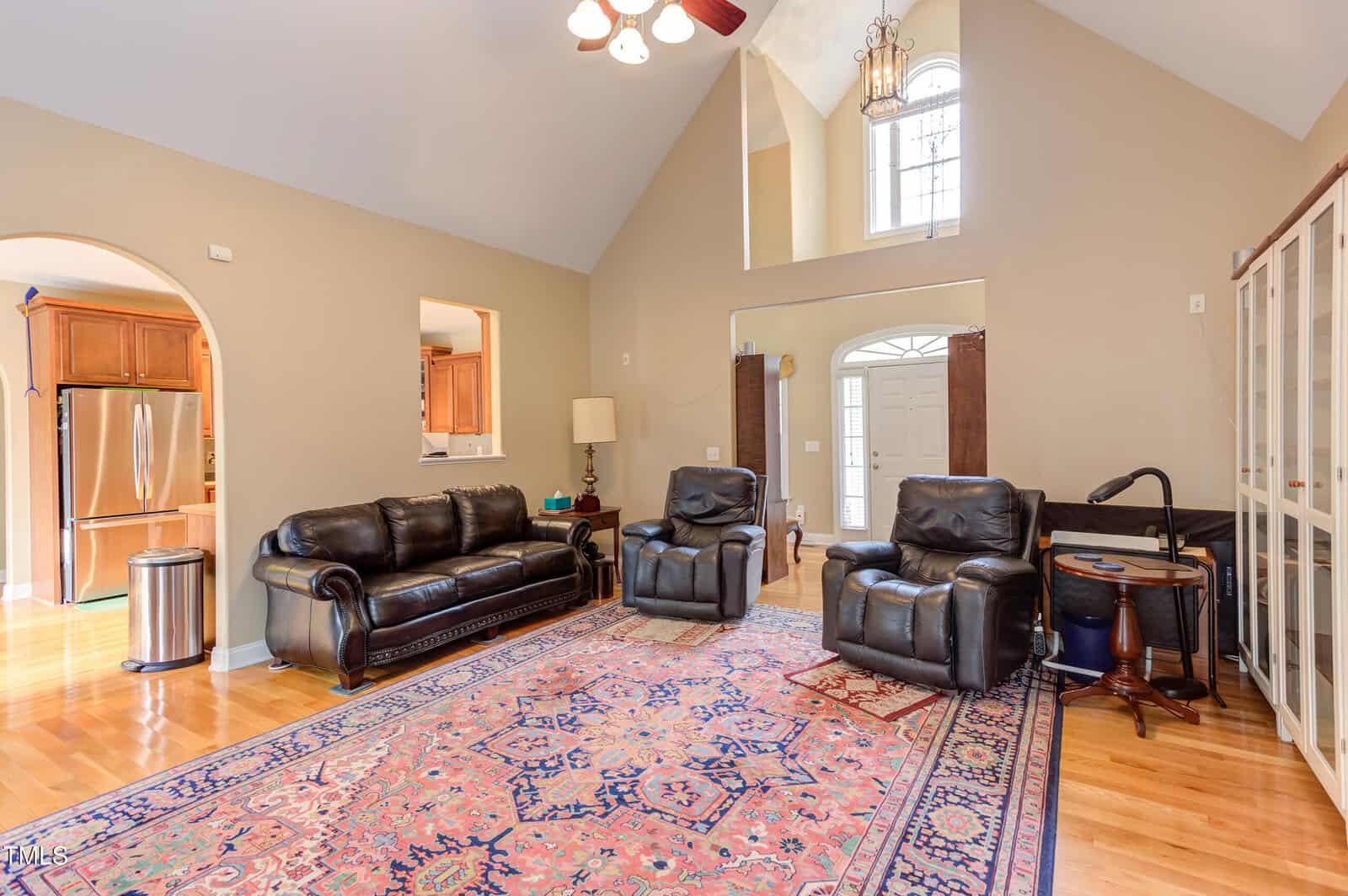 A living room with a high ceiling, leather sofa and armchairs, a patterned area rug, hardwood floors, and neutral walls. There's an archway to a kitchen, a large window above the front door, and a glass-fronted cabinet.