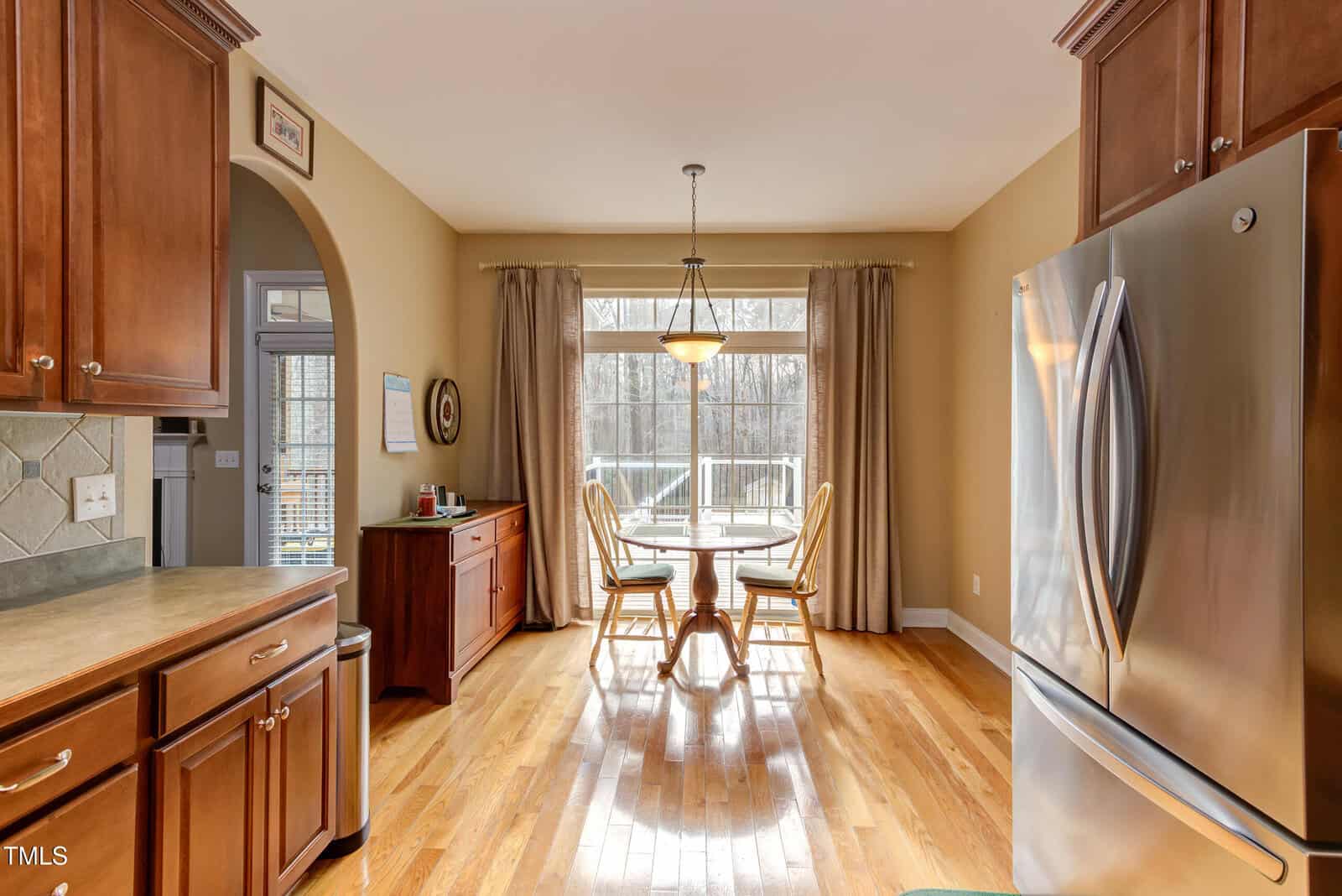 A bright kitchen with wooden cabinets and floors, a stainless steel refrigerator, and a dining area with a round table and four chairs next to large windows with beige curtains overlooking a deck and trees.