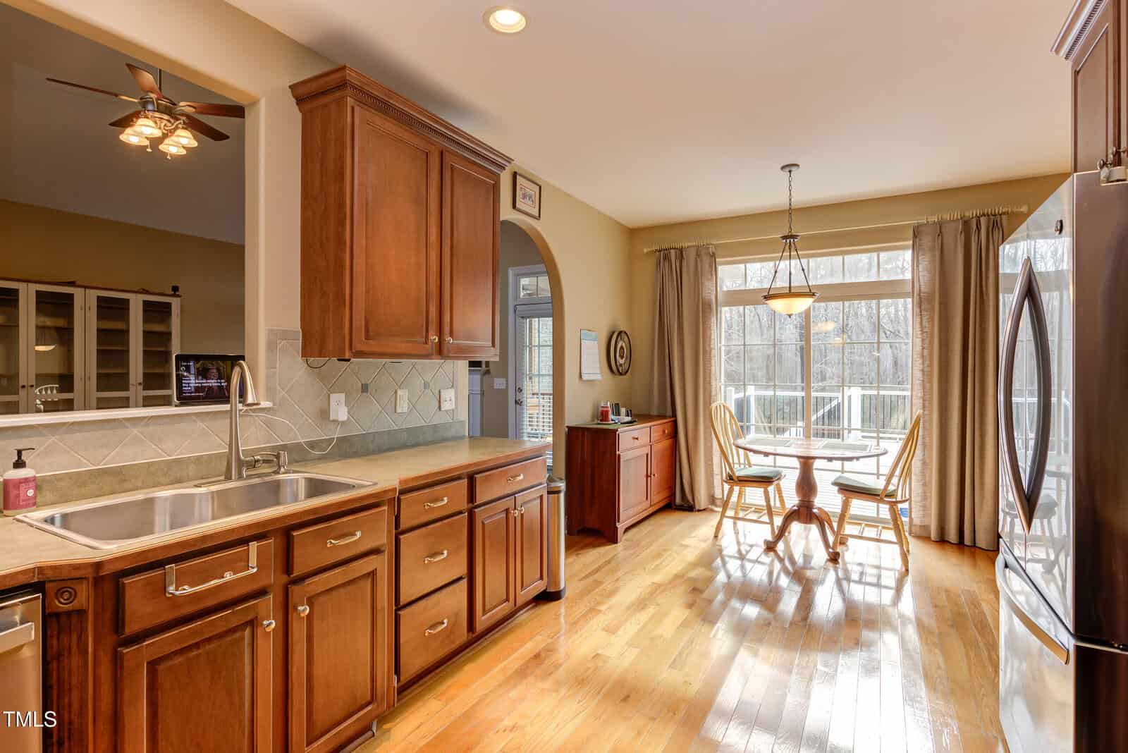 A bright kitchen with wooden cabinets, stainless steel appliances, a double sink, and a dining area with a round table and chairs by large windows overlooking a deck and wooded backyard.