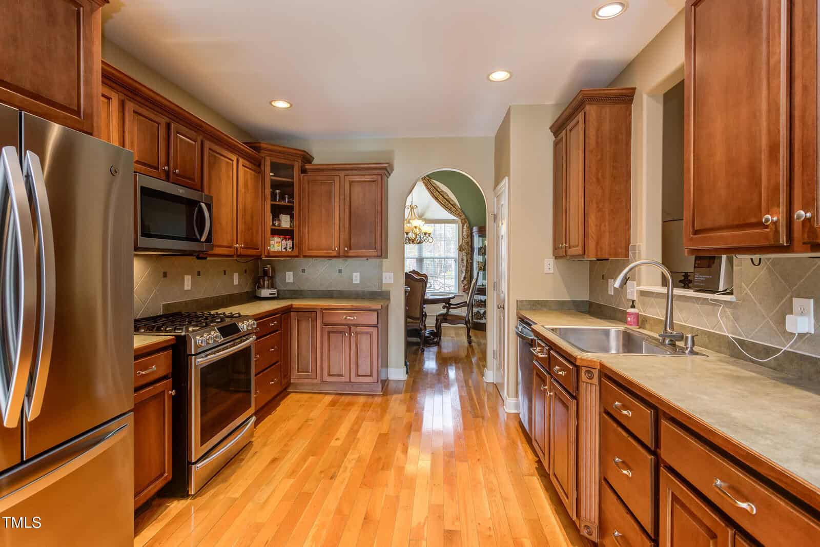 Spacious kitchen with wooden cabinets, stainless steel appliances, tile backsplash, and hardwood floors. A dining room with a chandelier is visible through an arched doorway.