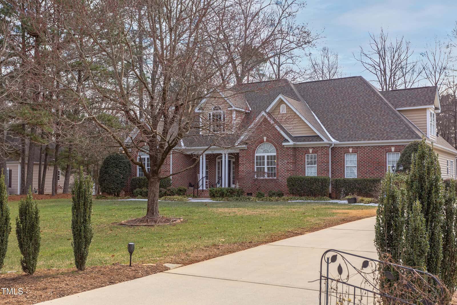 A two-story brick house with white trim, a covered front porch, and multiple gables, set on a spacious lawn with trees and shrubs. A long driveway leads to the front of the home.