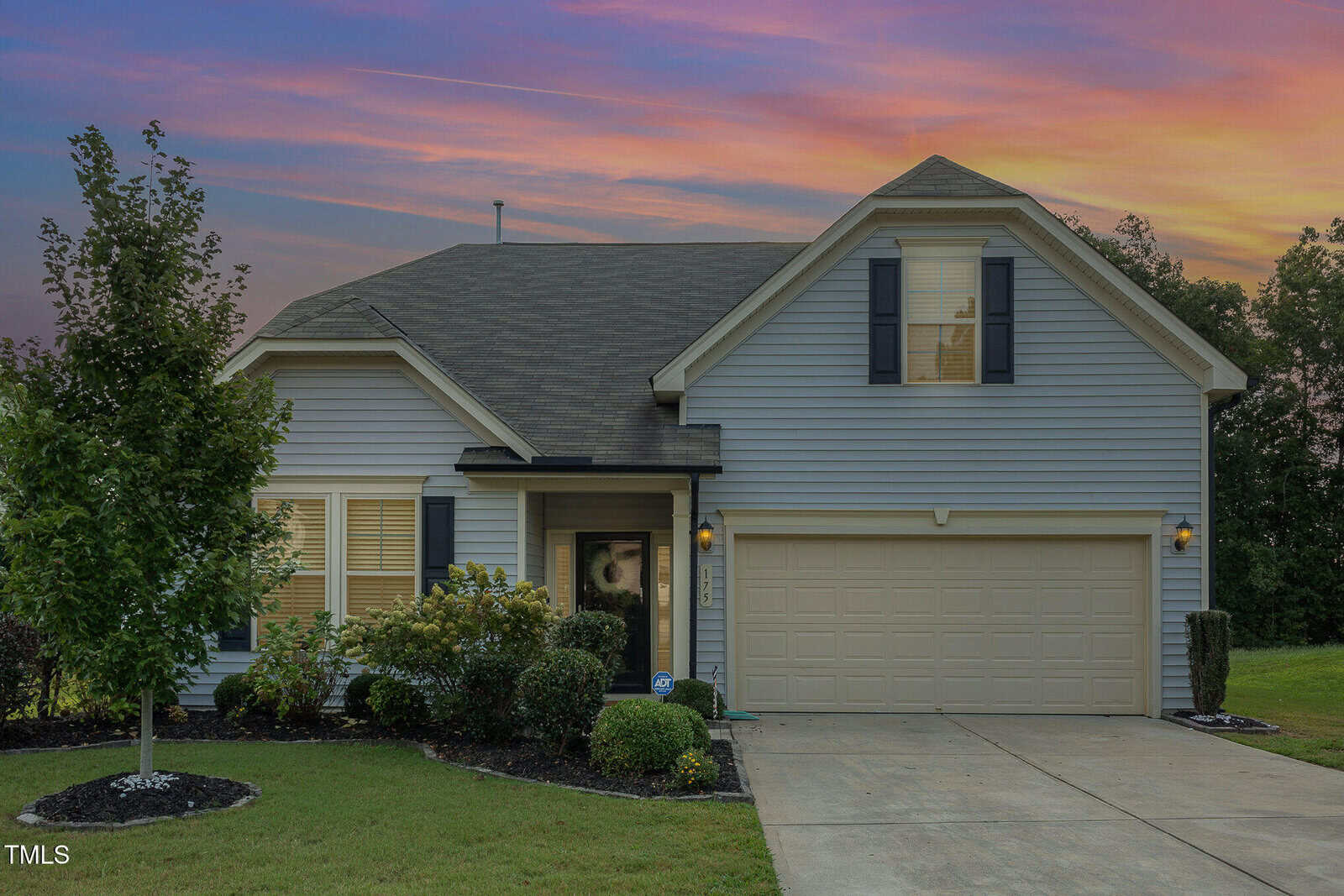 A two-story suburban house with gray siding, dark shutters, and a two-car garage is shown at sunset. The front yard features trimmed bushes, a tree, and a concrete driveway.
