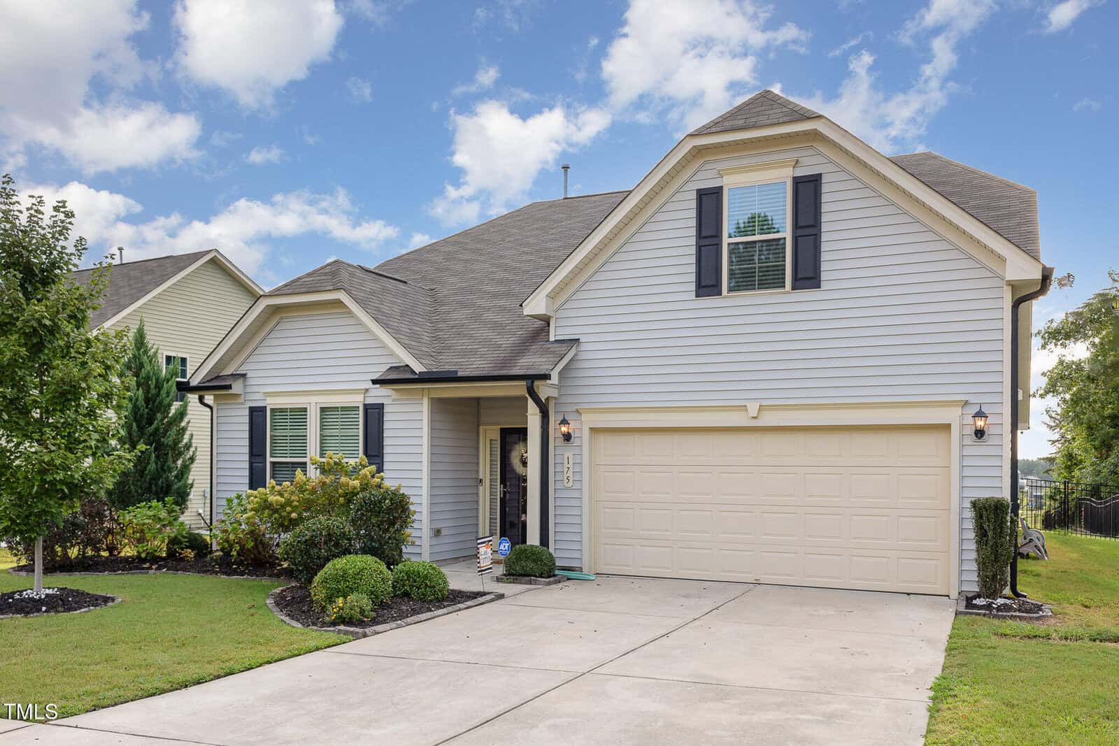 A two-story suburban house with light gray siding, black shutters, a double garage, a small covered entry, landscaped front yard, and a concrete driveway under a partly cloudy sky.