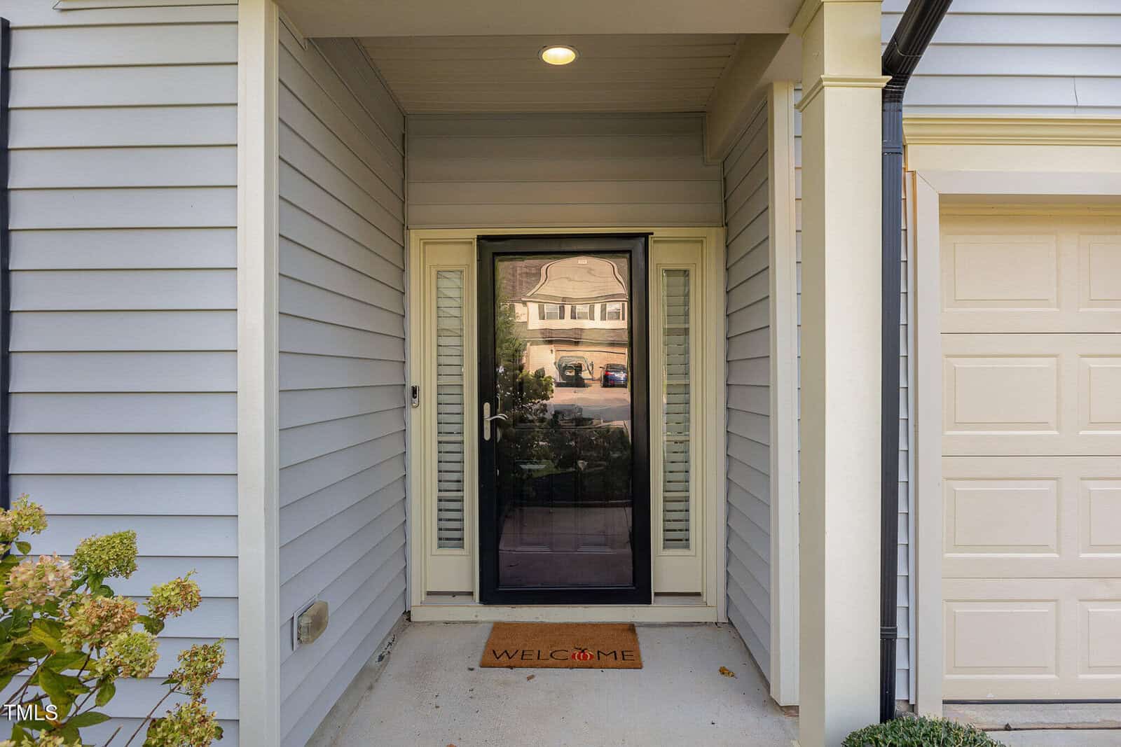 A front door with a glass storm door, beige siding, white trim, a “Welcome” doormat, and part of a garage door visible on the right. Bushes and a porch light are also visible.