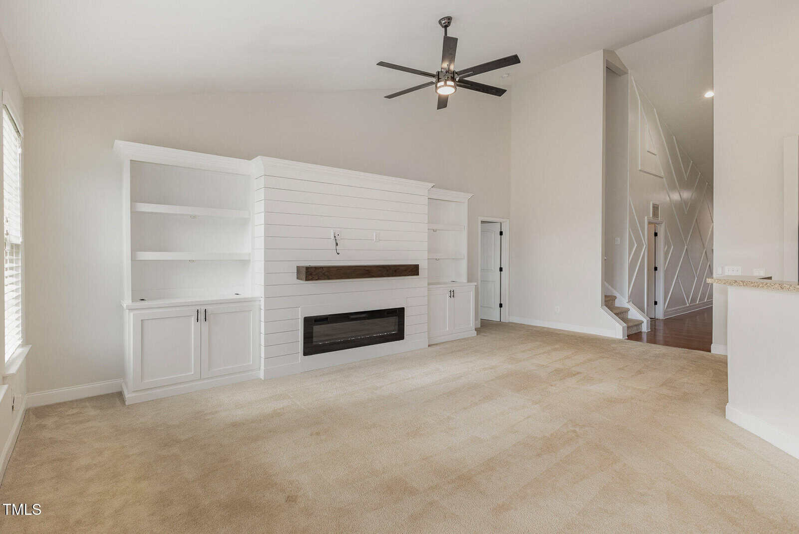 A spacious living room with beige carpet, white built-in shelves and cabinets, a modern fireplace, a ceiling fan, and a staircase visible in the background. The walls are painted light beige.