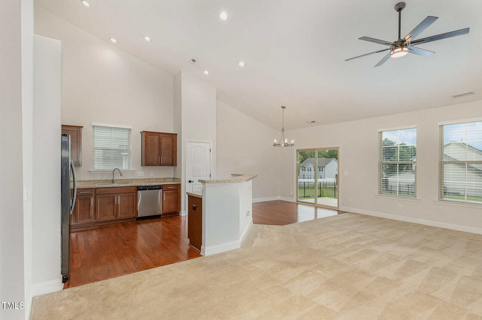 Open-concept living space with a kitchen featuring wood cabinets and stainless steel appliances, a carpeted living area, dining nook with chandelier, large windows, and ceiling fan. Bright, neutral colors throughout.