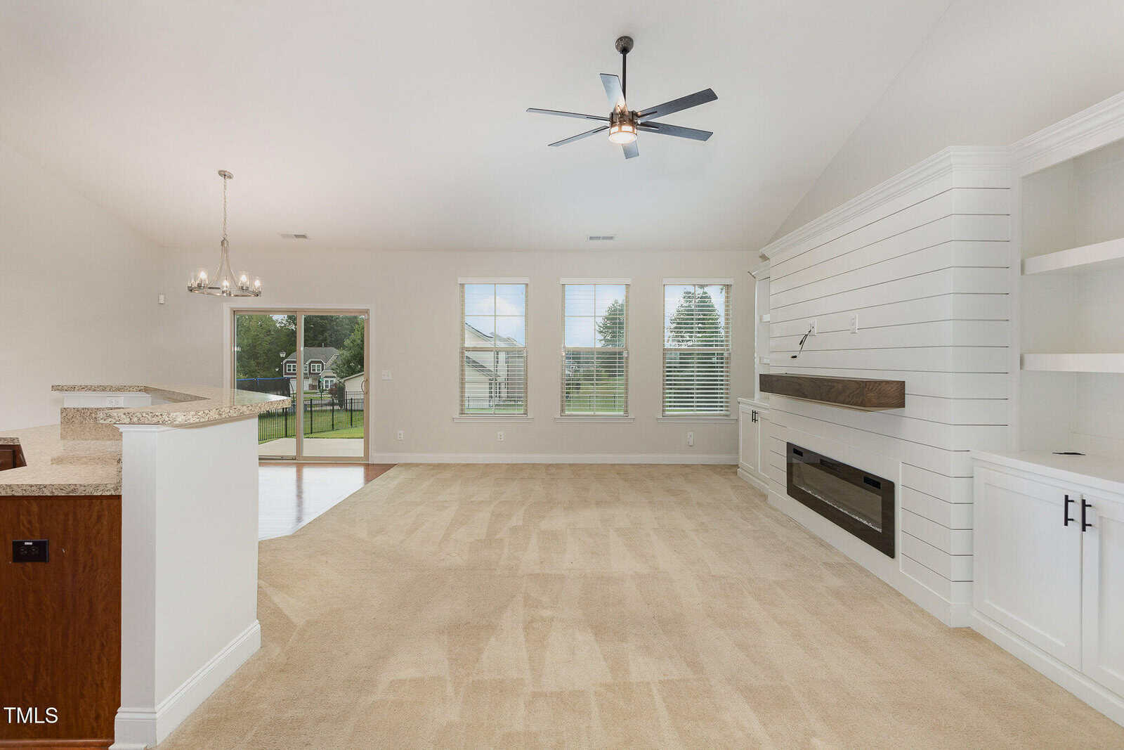 Spacious living room with beige carpet, large windows, built-in shelves, white shiplap accent wall, ceiling fan, fireplace, and open view to the kitchen and dining area; backyard visible through glass door.