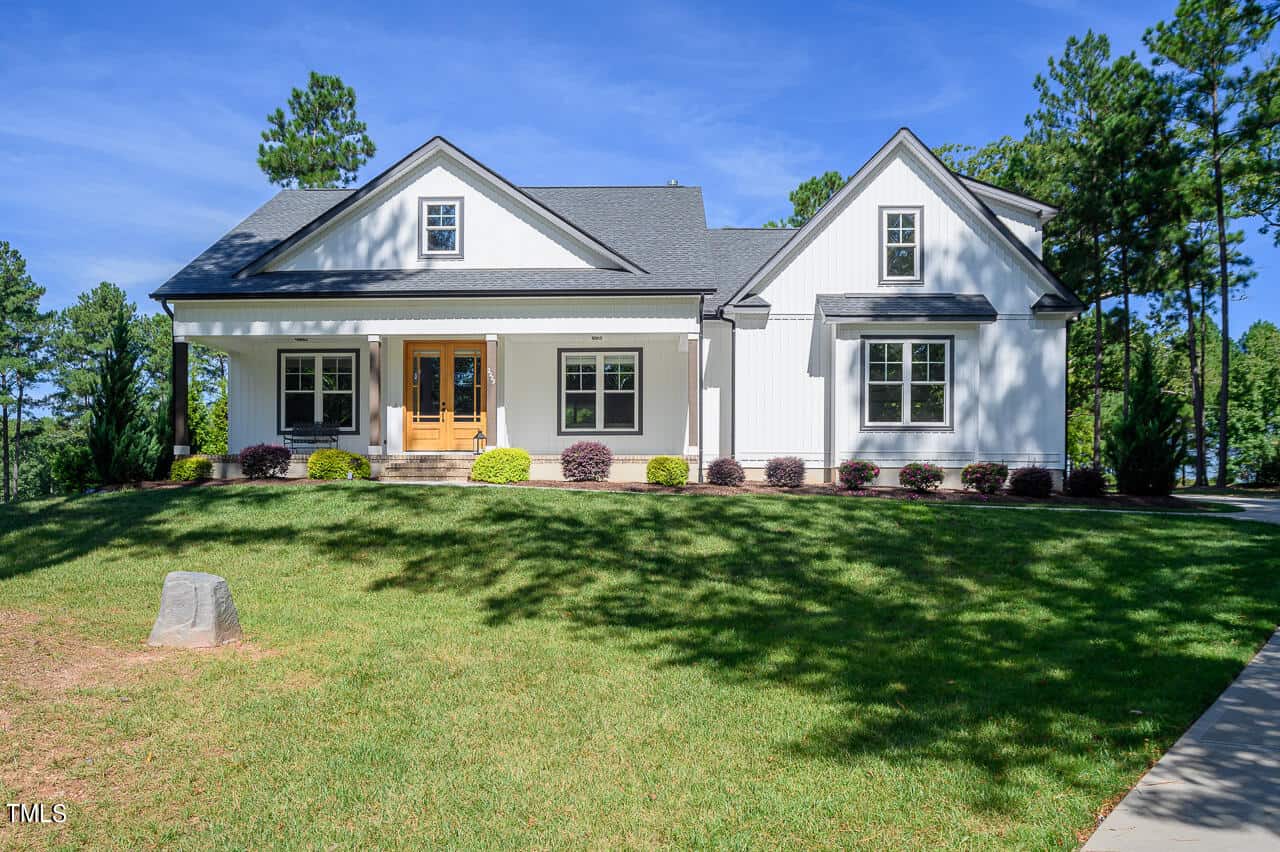 A two-story white house at 2223 Grove Hill Road in Franklinton boasts a dark gray roof set against a backdrop of trees. It features a wooden front door, a porch with white railings, and well-maintained landscaping. The expansive lawn unfolds beneath a clear, blue sky.