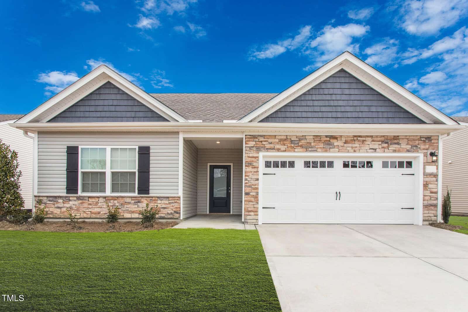 Single-story suburban house with gray siding, stone accents, and black shutters. Features a double garage with white doors, a small covered entryway, and a manicured lawn under a blue sky with scattered clouds.