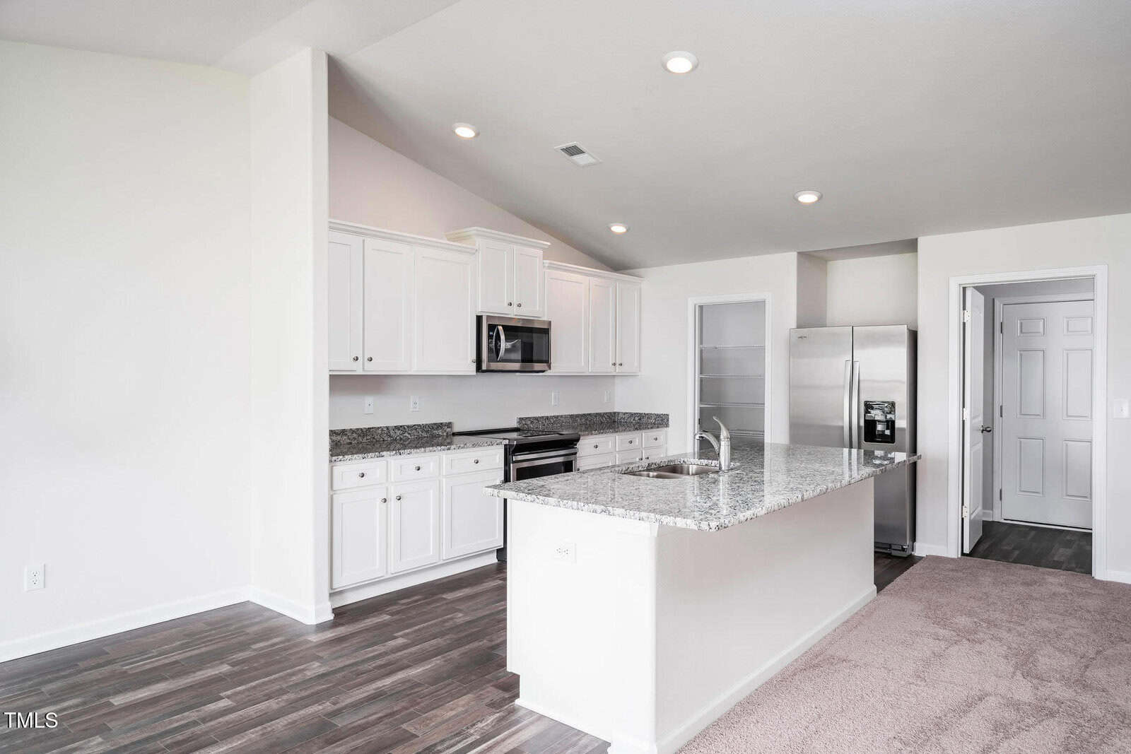 A modern kitchen with white cabinets, granite countertops, stainless steel appliances, and wood flooring. The kitchen island includes a sink, and the adjacent area is carpeted. Walls are painted white.