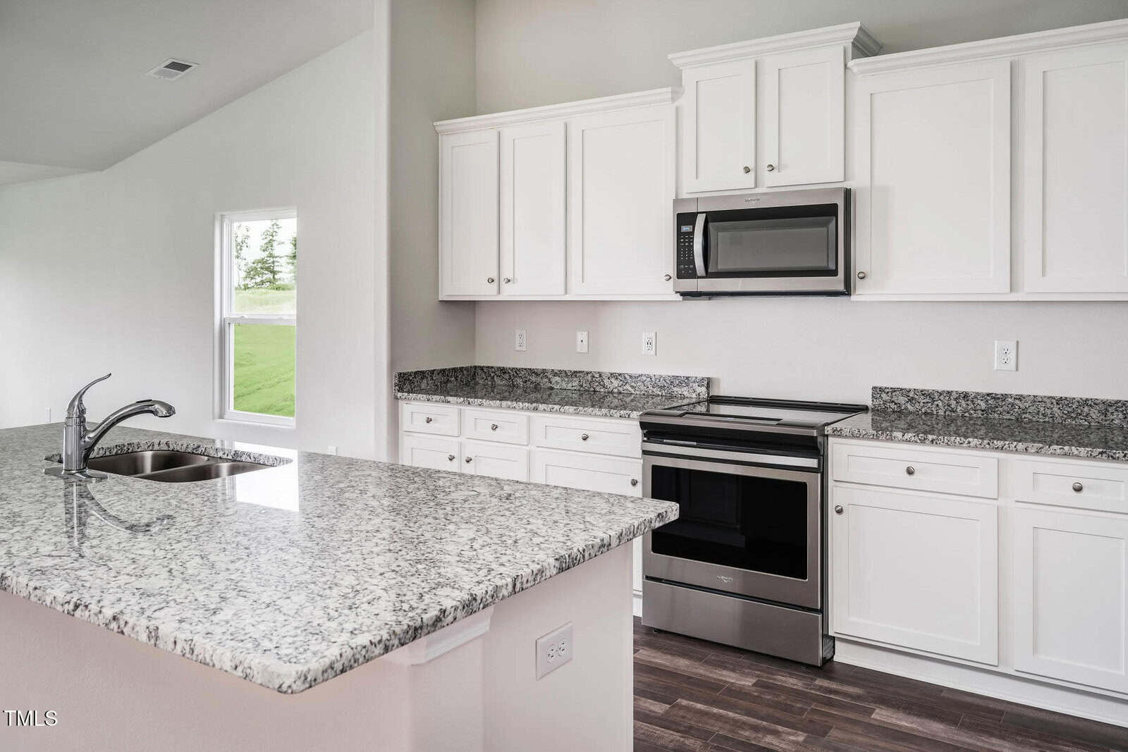 A modern kitchen with white cabinets, stainless steel appliances, granite countertops, a double sink with a chrome faucet, and a window providing natural light. The floor is dark wood.