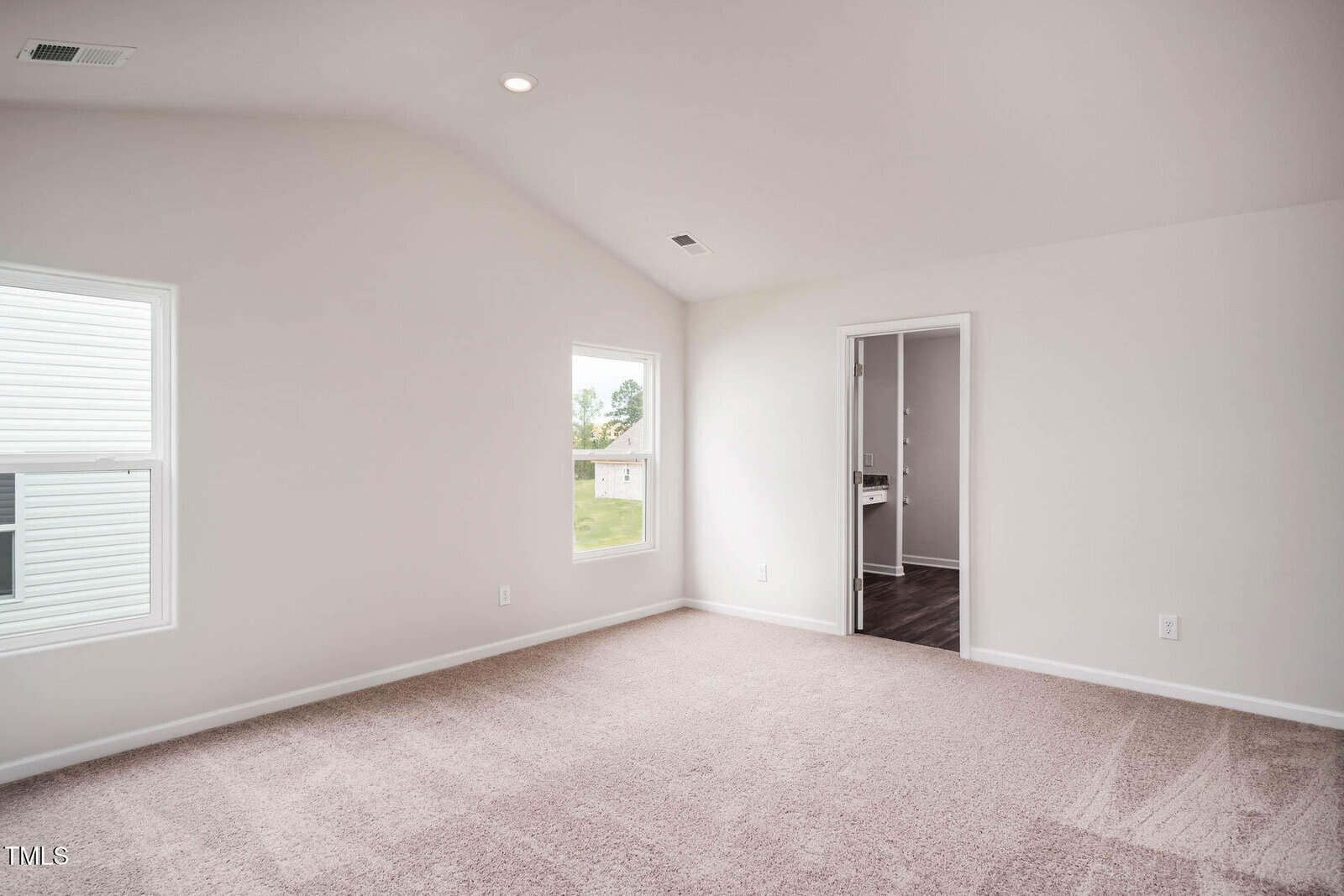 A bright, empty room with beige carpet, white walls, two windows, a vent in the ceiling, and a door leading to an adjoining room with dark flooring visible.
