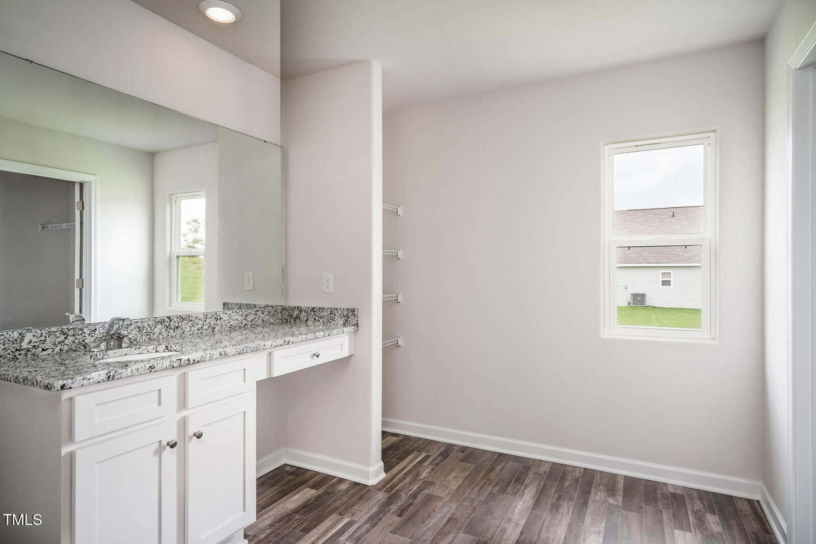 Bathroom with white cabinets, granite countertop, large wall mirror, wood-look flooring, towel shelves, and two windows providing natural light. The space is empty, and the walls are painted a light color.