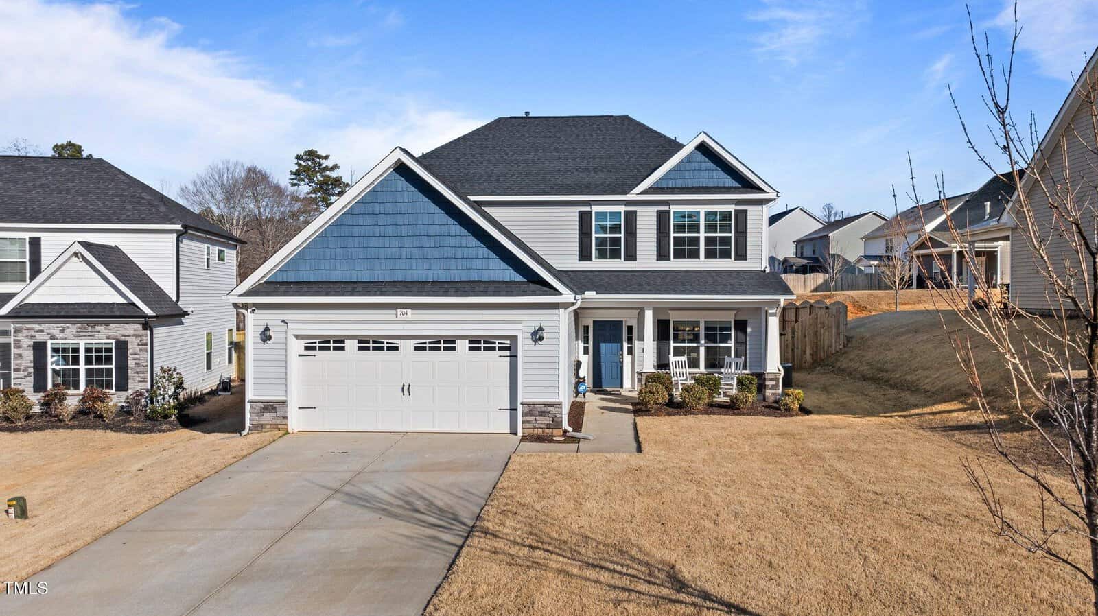 A two-story suburban house with blue and white siding, a covered front porch, and a two-car garage. The house is surrounded by a dry, brown lawn and neighboring houses are visible on both sides.